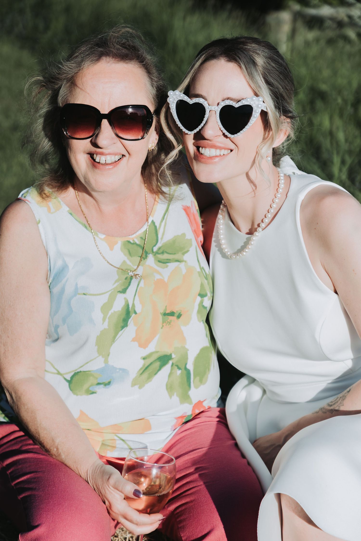 Two people wearing sunglasses and light colored tops sitting together outdoors on a sunny day.