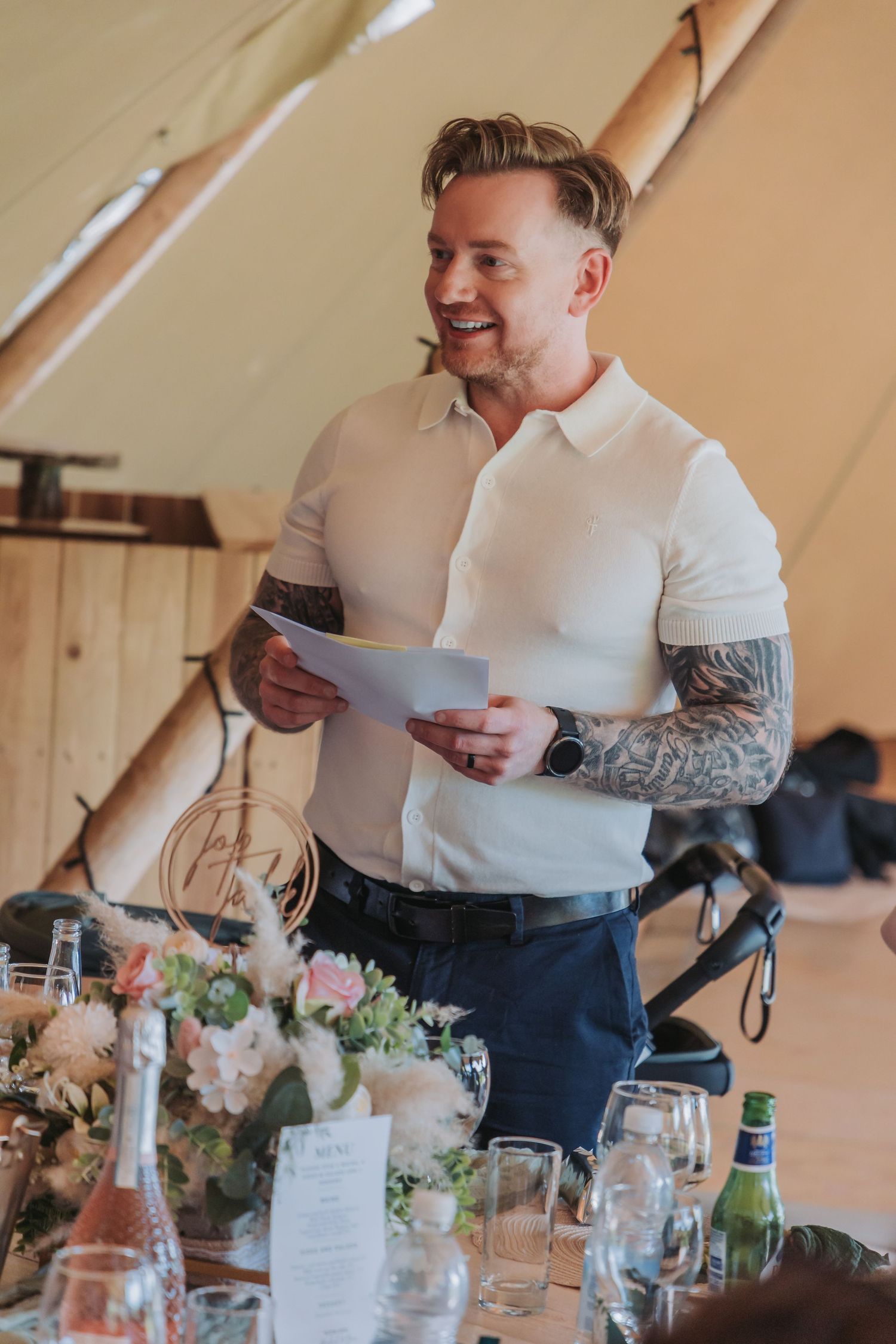 A person in a white shirt reads from papers while standing at a decorated table with floral arrangements and glassware.