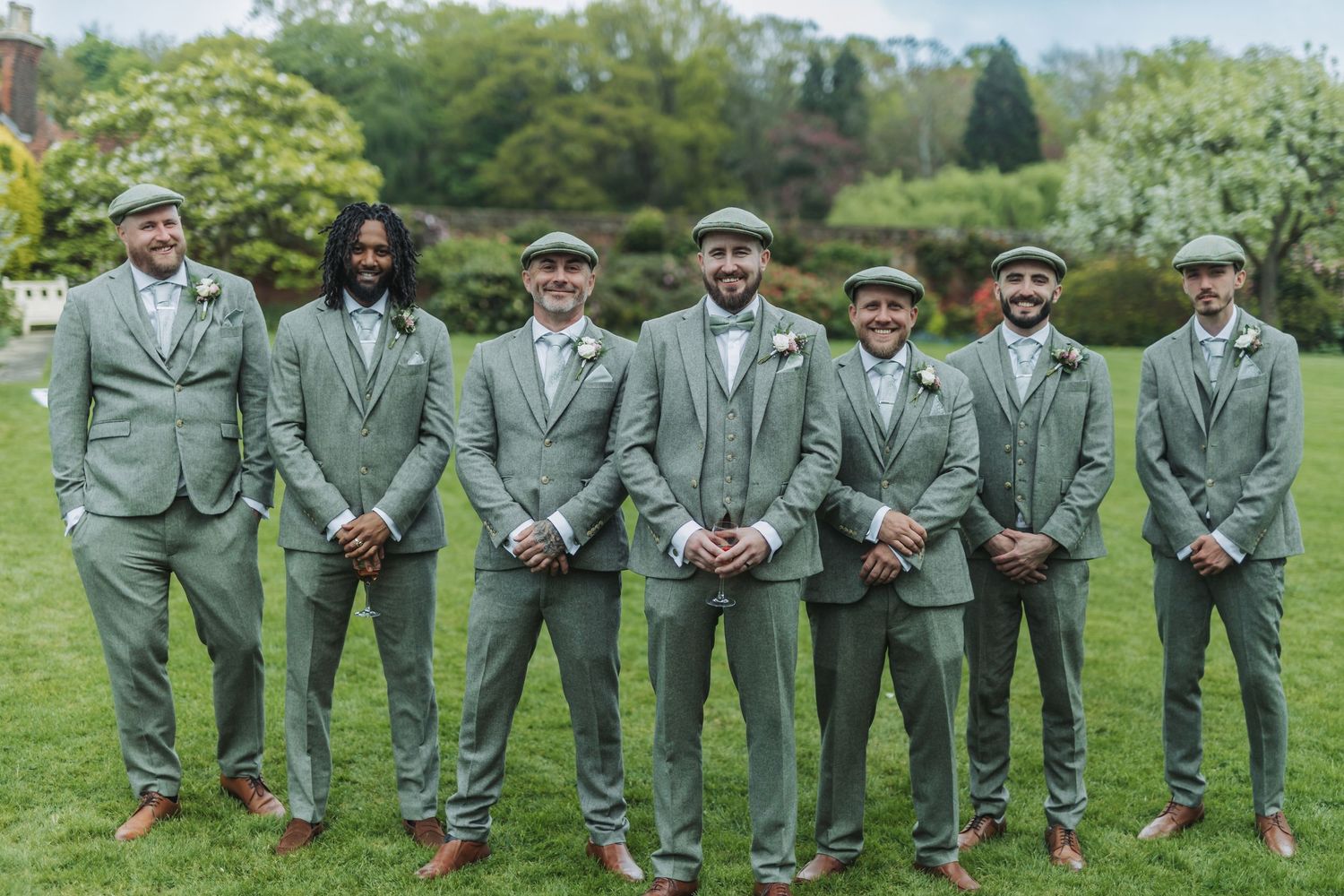 Wedding party in sage green suits poses together on lush grass for outdoor group photos.