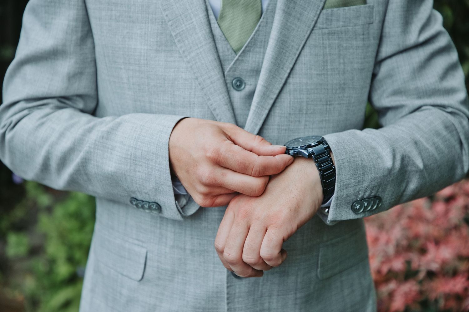 Man in gray three-piece suit adjusting his watch, wearing a sage green tie and pocket square.