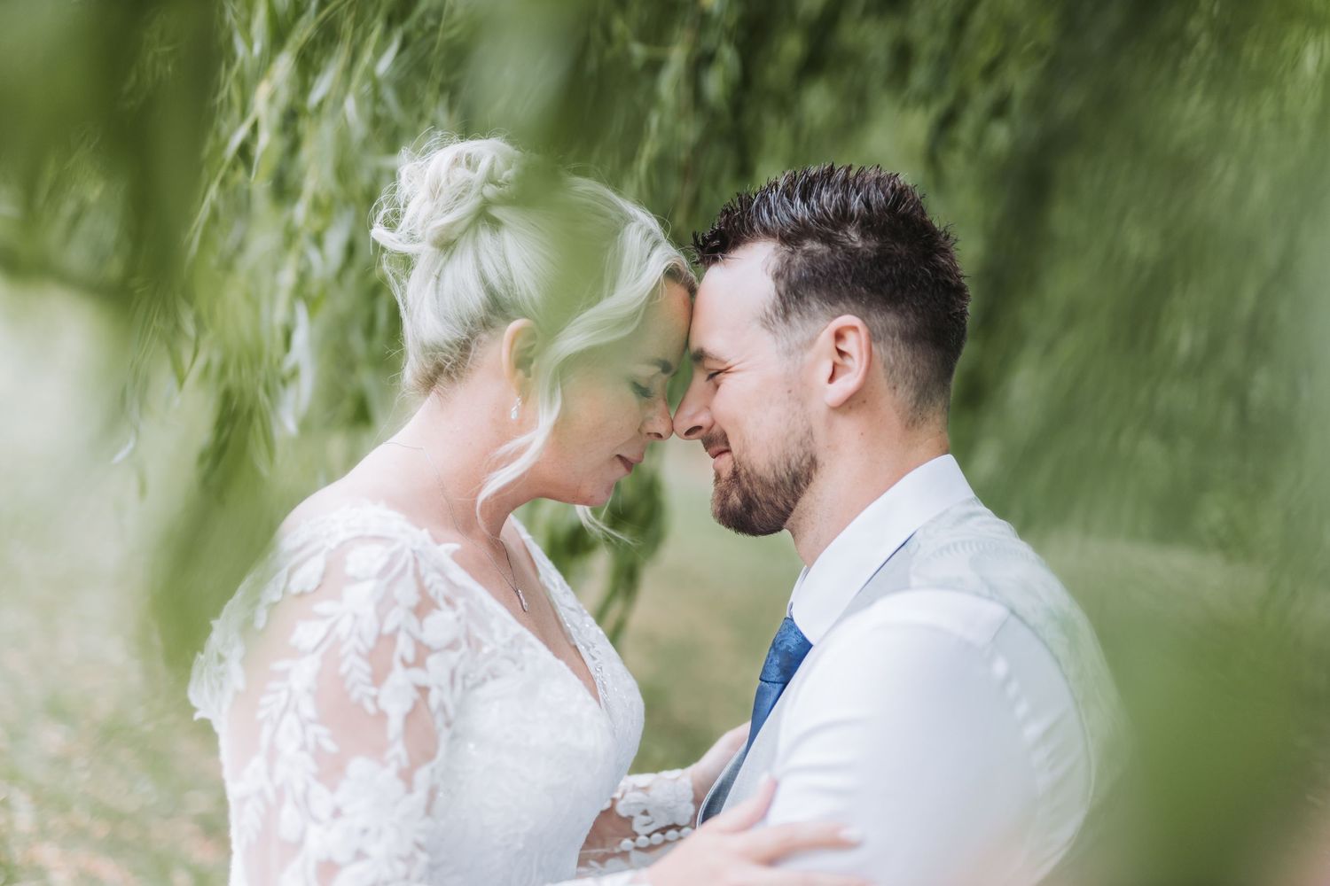 A romantic wedding couple shares an intimate moment together in a dreamy outdoor garden setting with soft green foliage.