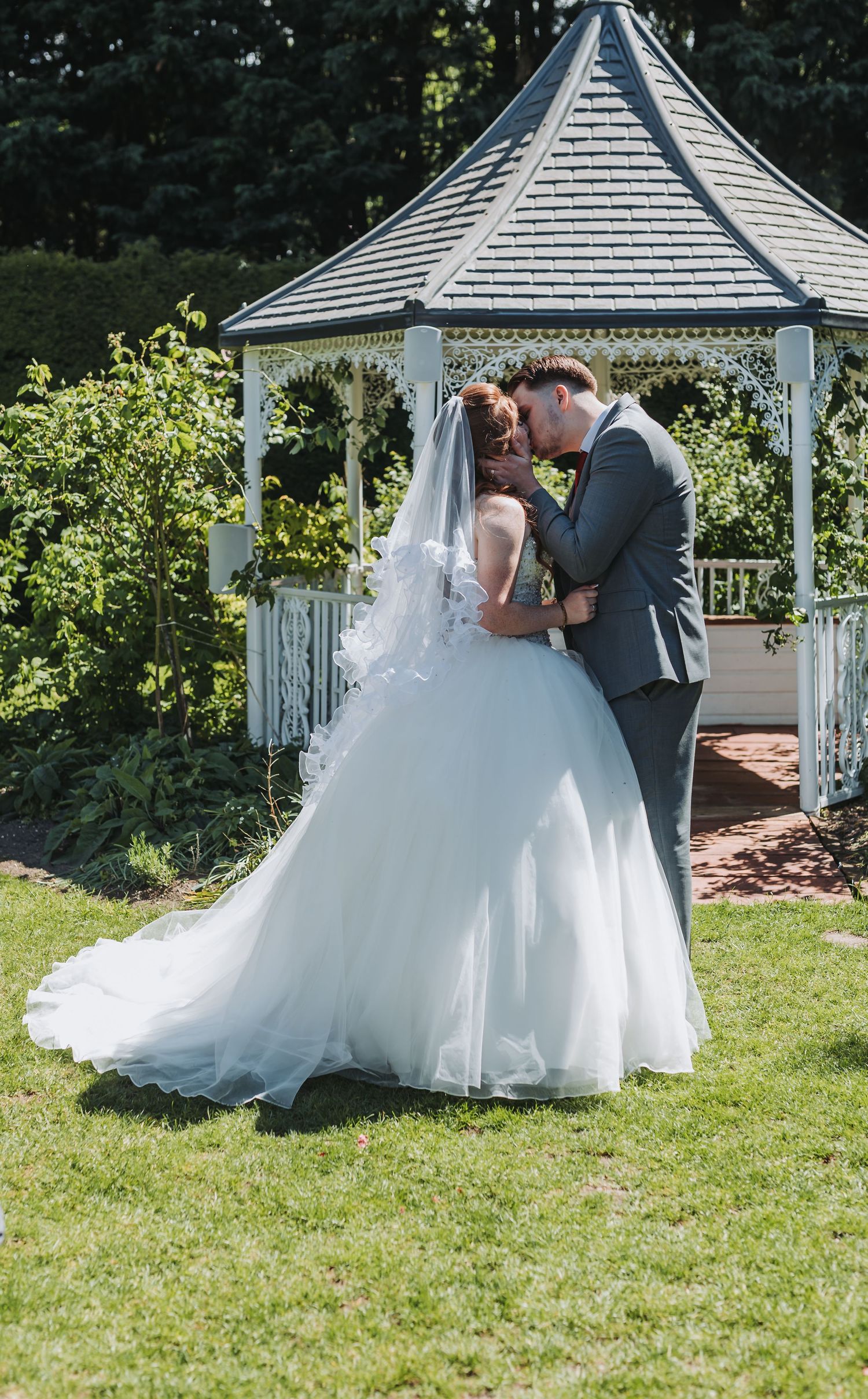 A romantic wedding moment captured at a white garden gazebo with a bride in a flowing tulle gown and long veil.