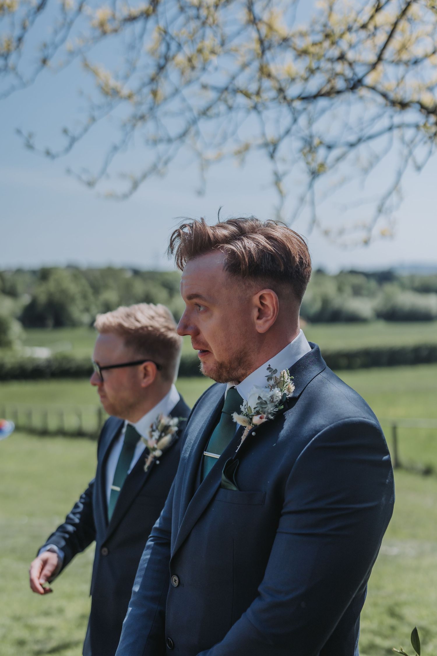 Three groomsmen in navy suits with floral boutonnieres stand together in a sunny outdoor field setting.