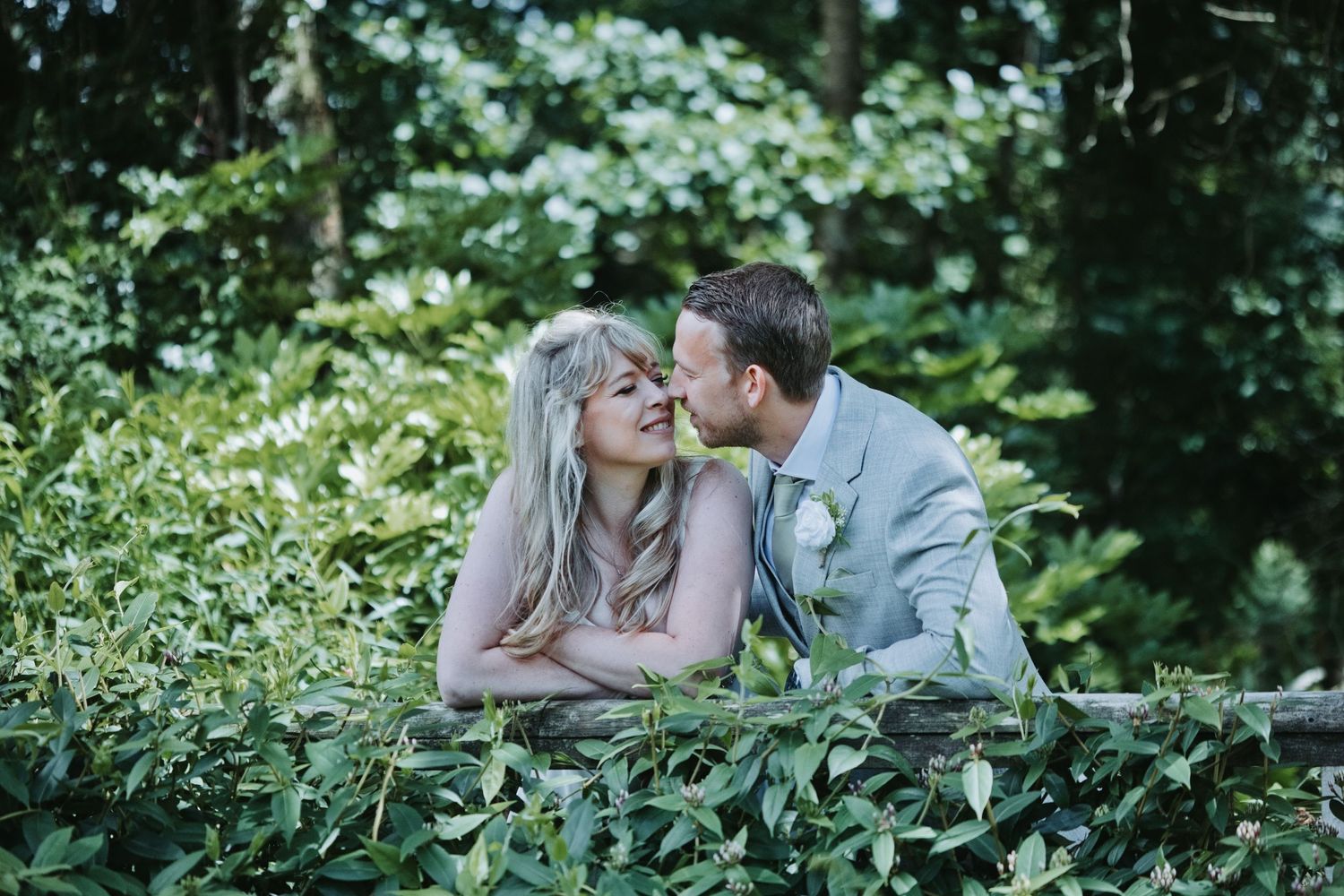 A bride and groom share a romantic kiss surrounded by lush green foliage in an outdoor garden wedding setting.