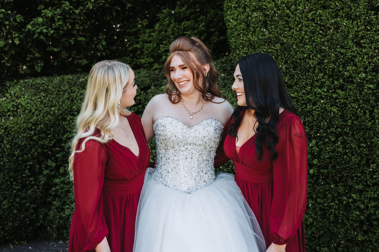 Bridesmaids in burgundy dresses gather around a bride in a white lace gown against a green hedge backdrop.