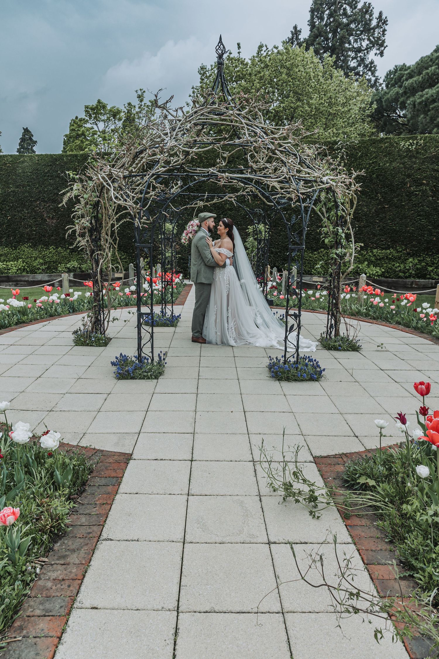 Wedding ceremony setup in a garden with brick pathways, white archway, and blooming red flowers in a modern courtyard setting.