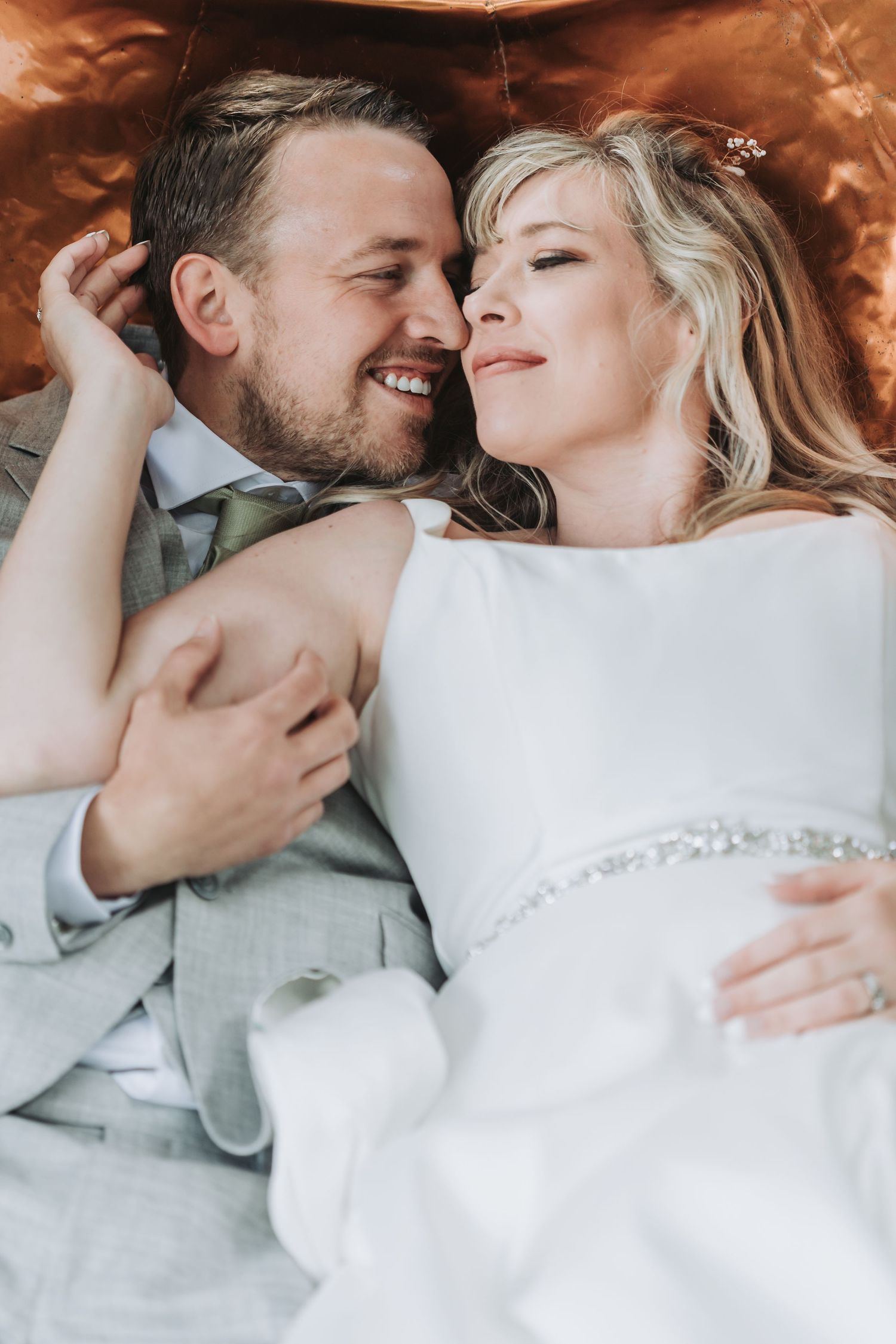 A smiling bride and groom in wedding attire lying together, sharing a tender romantic moment on their wedding day.