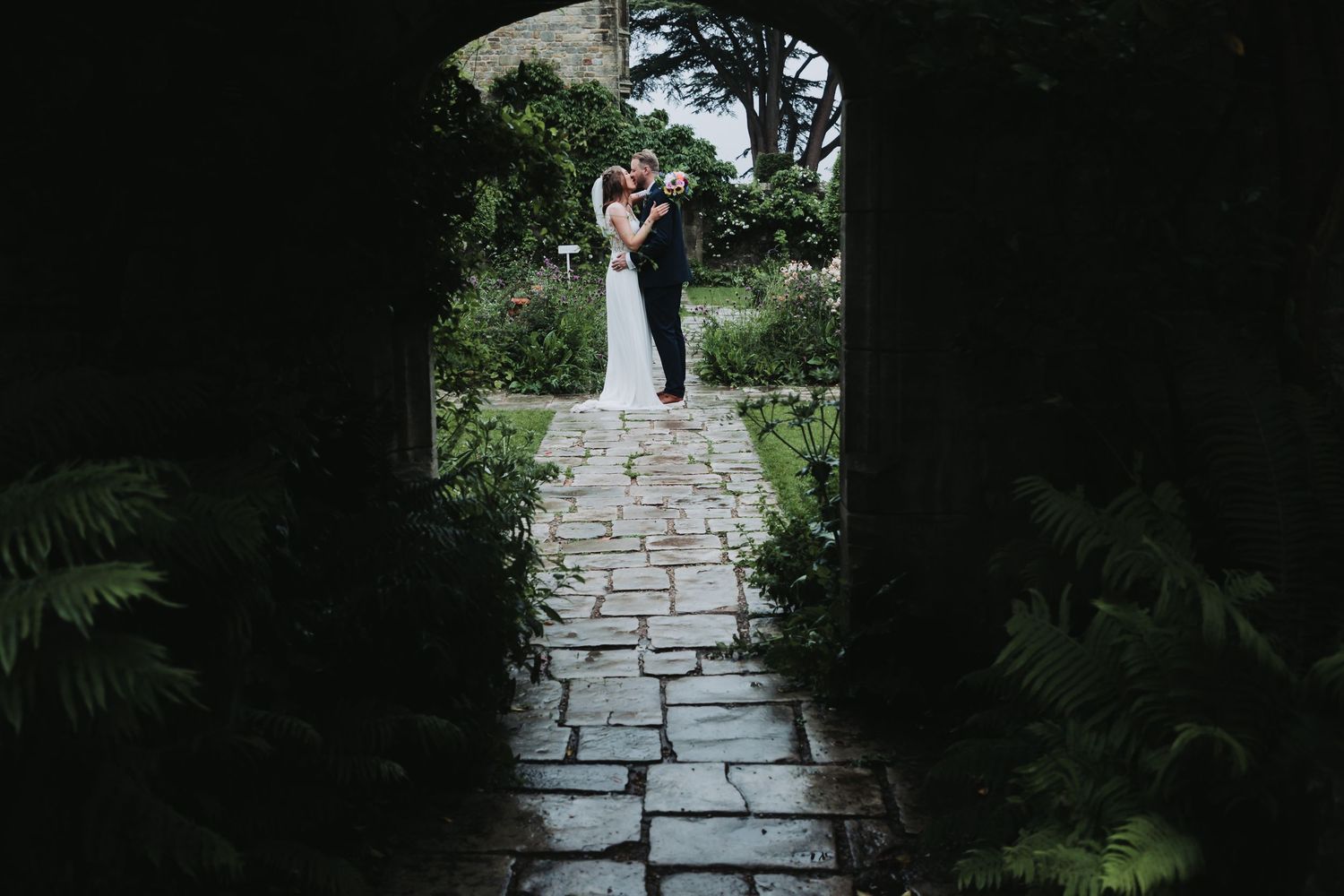 Couple embracing in a lush garden framed by a stone archway on a cobblestone path, perfect for a romantic wedding setting.