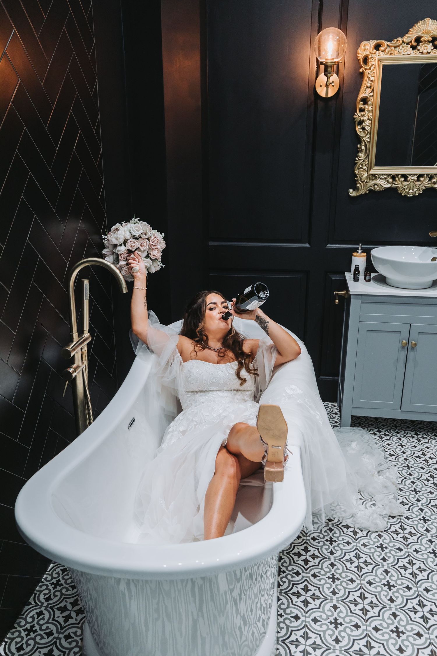 A person in a white wedding dress sits in a luxurious clawfoot bathtub while drinking wine in an elegant bathroom.