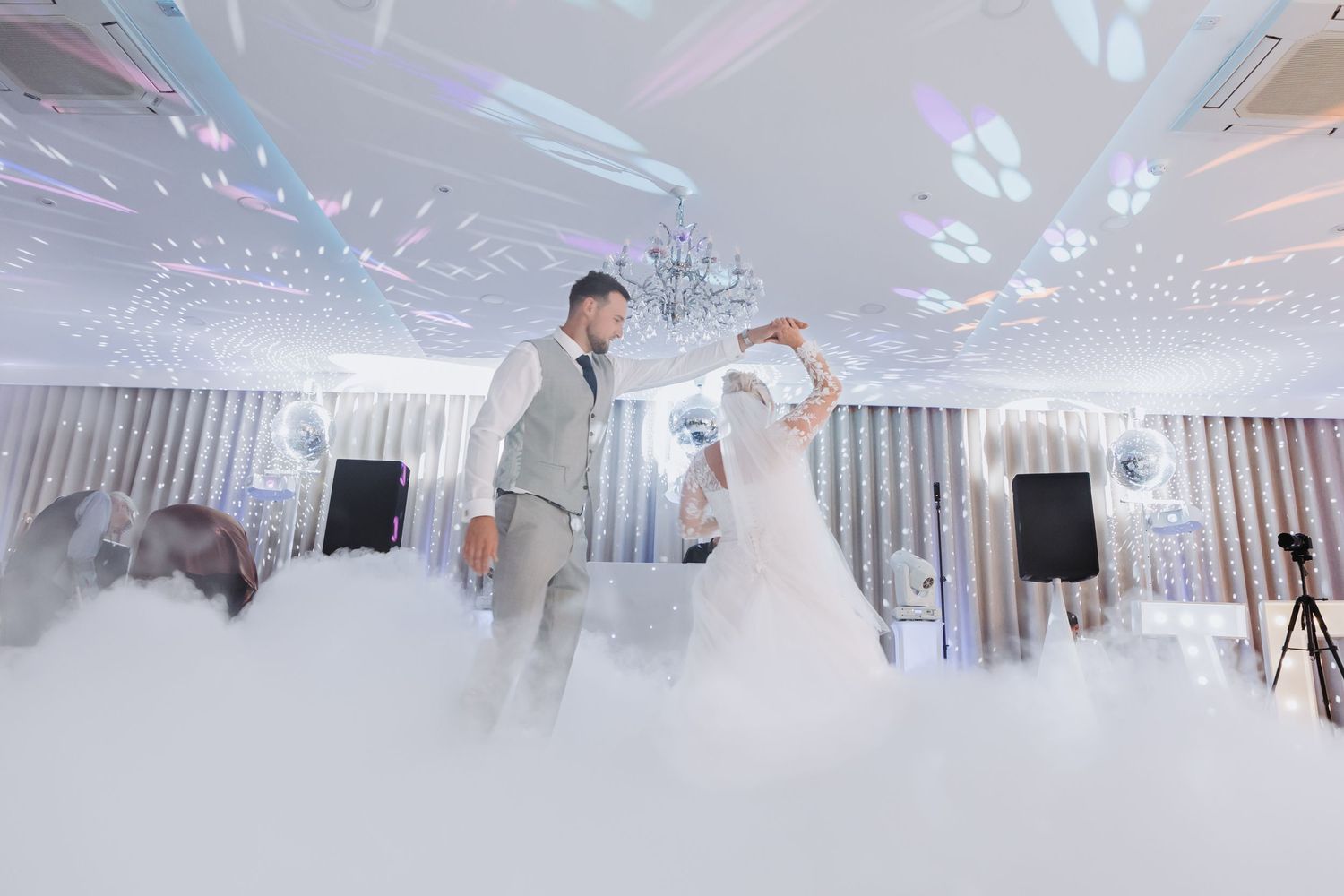 Couple dancing their first dance on a misty white dance floor with dramatic lighting and bubbles at an elegant wedding reception.