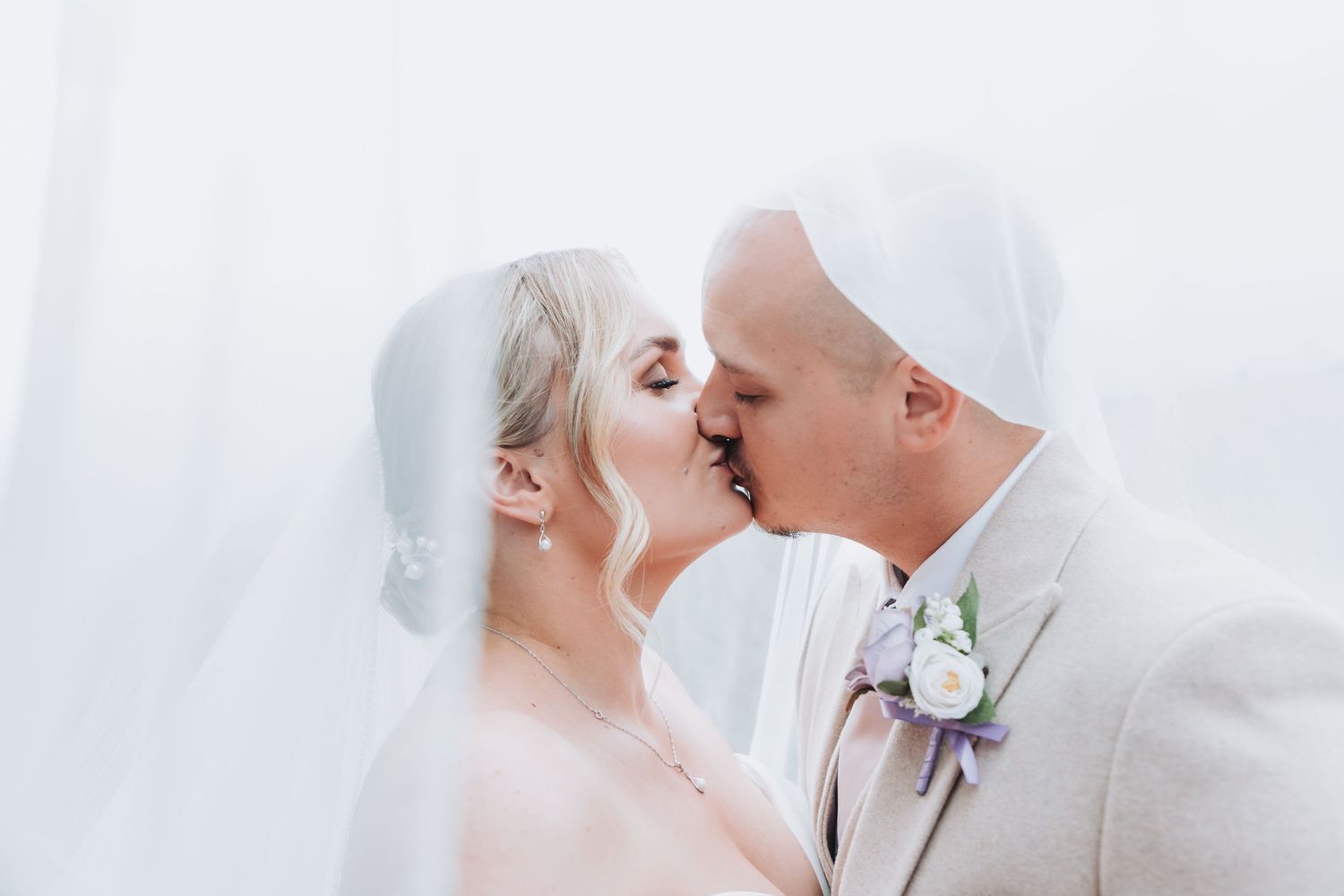 A tender wedding kiss captured through a soft white veil with a purple boutonniere visible on the groom's lapel.
