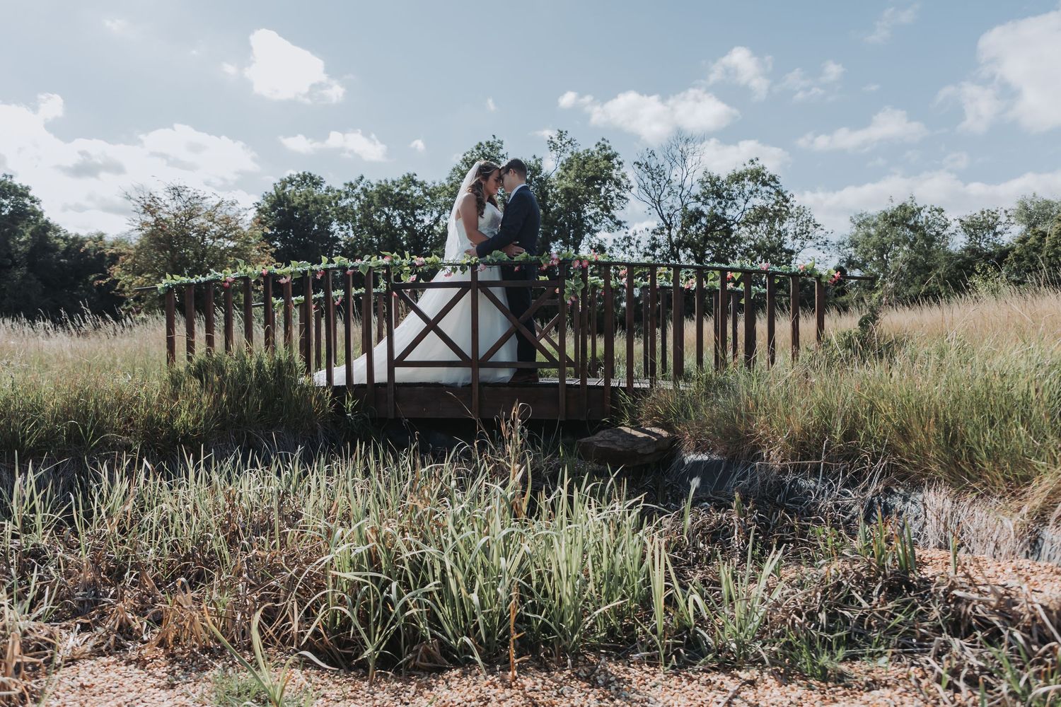 A panoramic view of a serene wetland landscape with tall grass, trees, and a wooden structure under a blue sky.