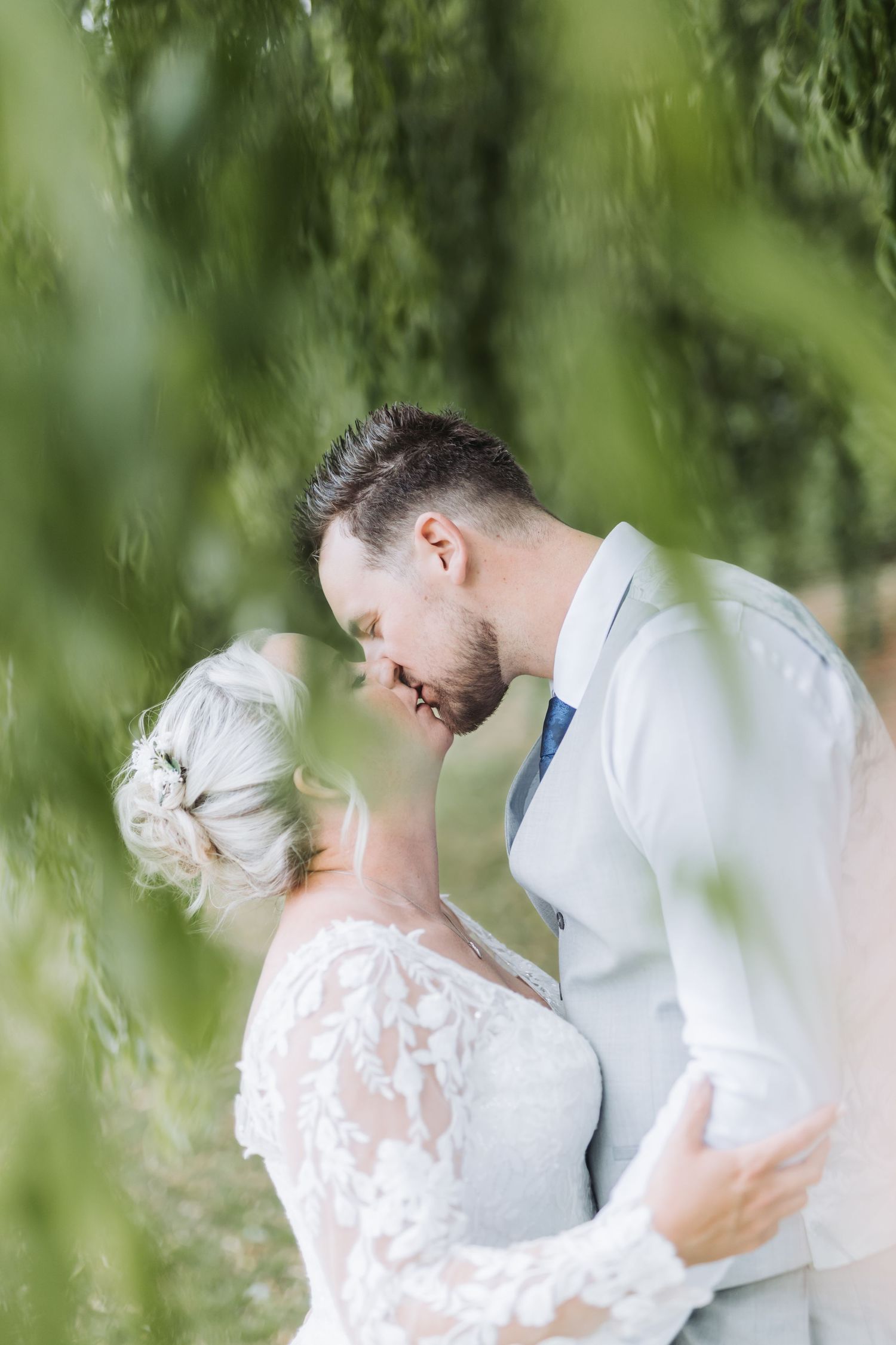 A romantic wedding couple embraces under cascading willow tree branches in an intimate outdoor setting.