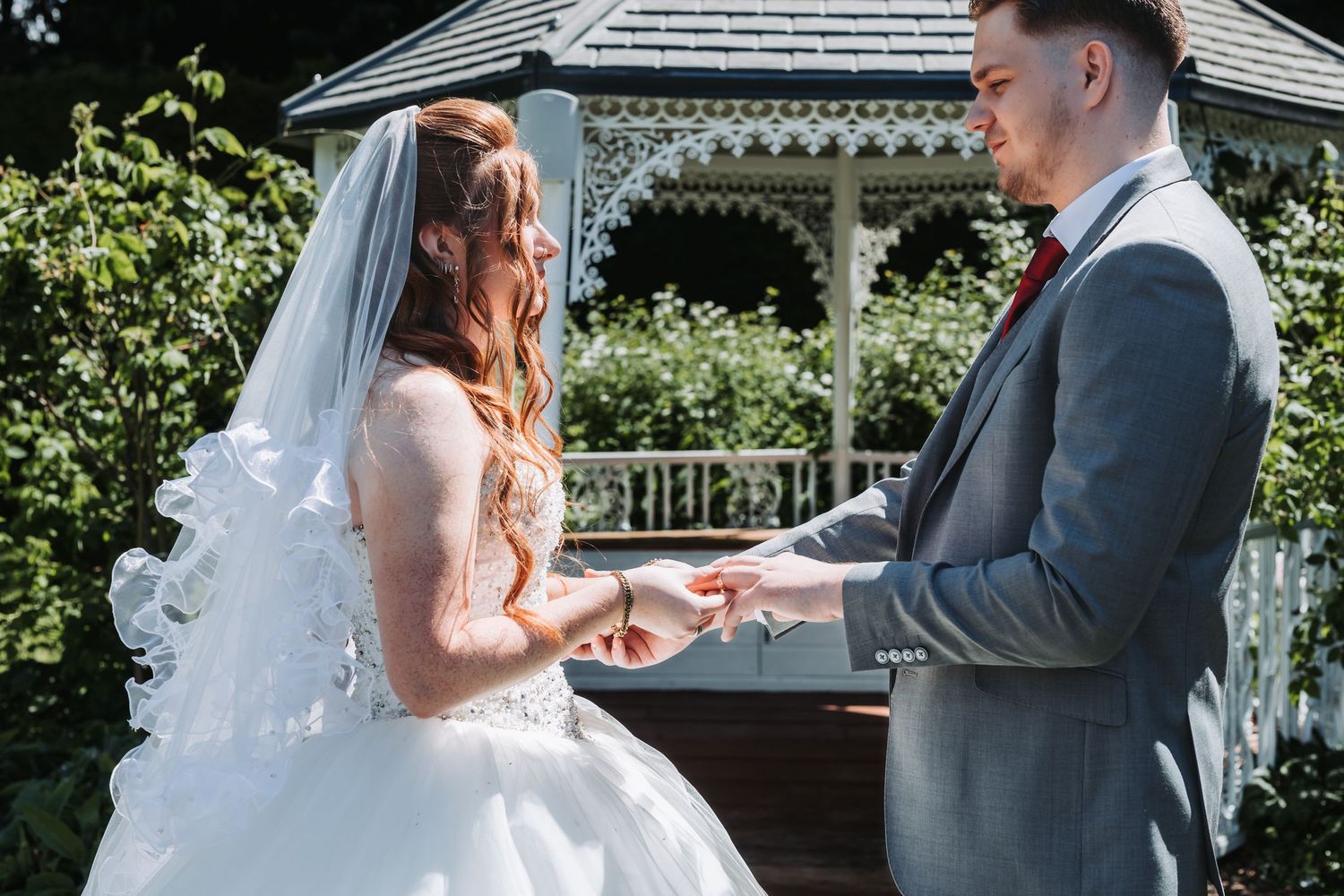 A couple exchanges rings during an outdoor wedding ceremony in front of a white gazebo on a sunny day.