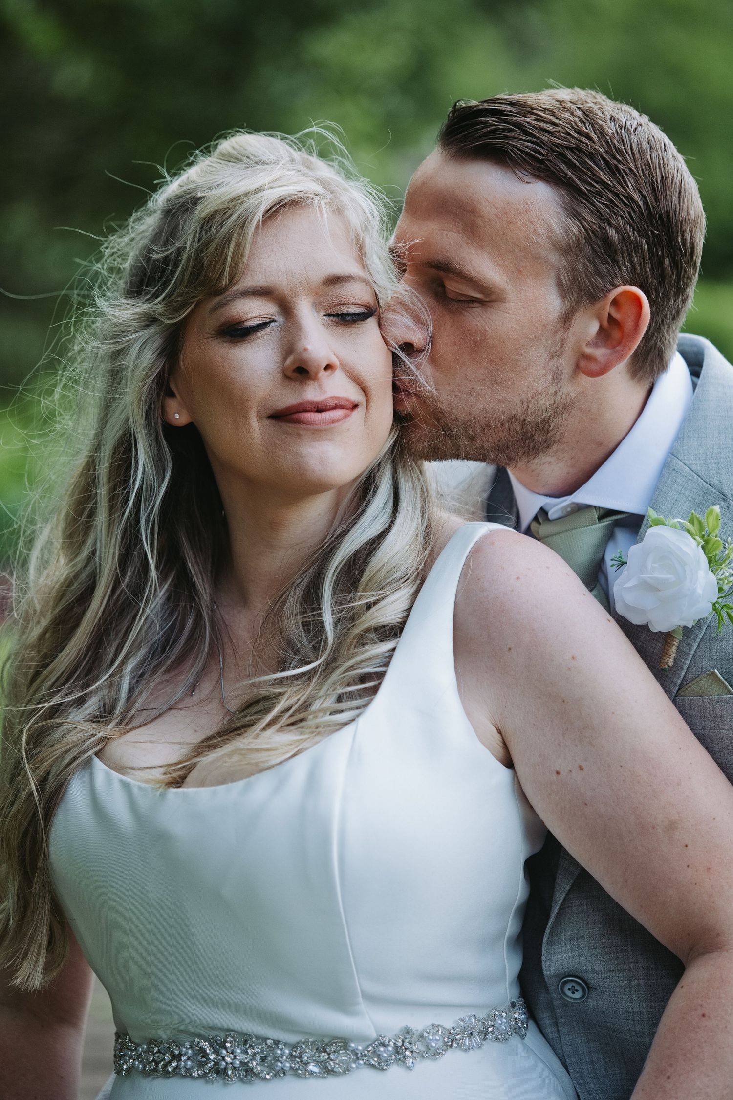 Groom in gray suit kissing bride on cheek as she smiles with eyes closed in white wedding dress outdoors.