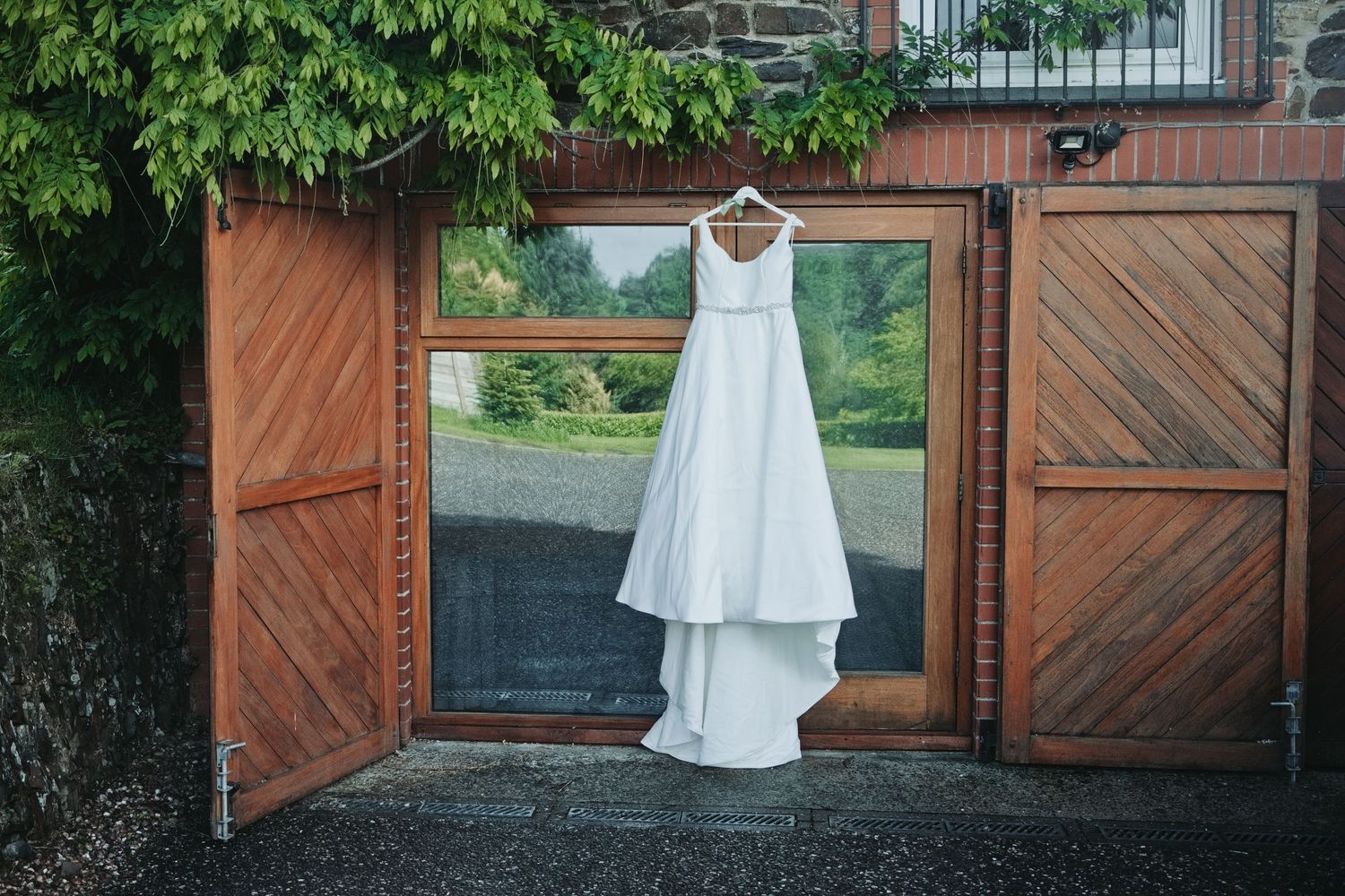 White wedding dress hanging on wooden barn doors with ivy-covered stone wall backdrop.