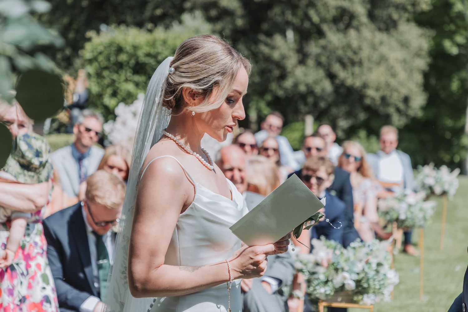 A couple exchanges wedding rings during an outdoor garden ceremony surrounded by guests and floral decorations.