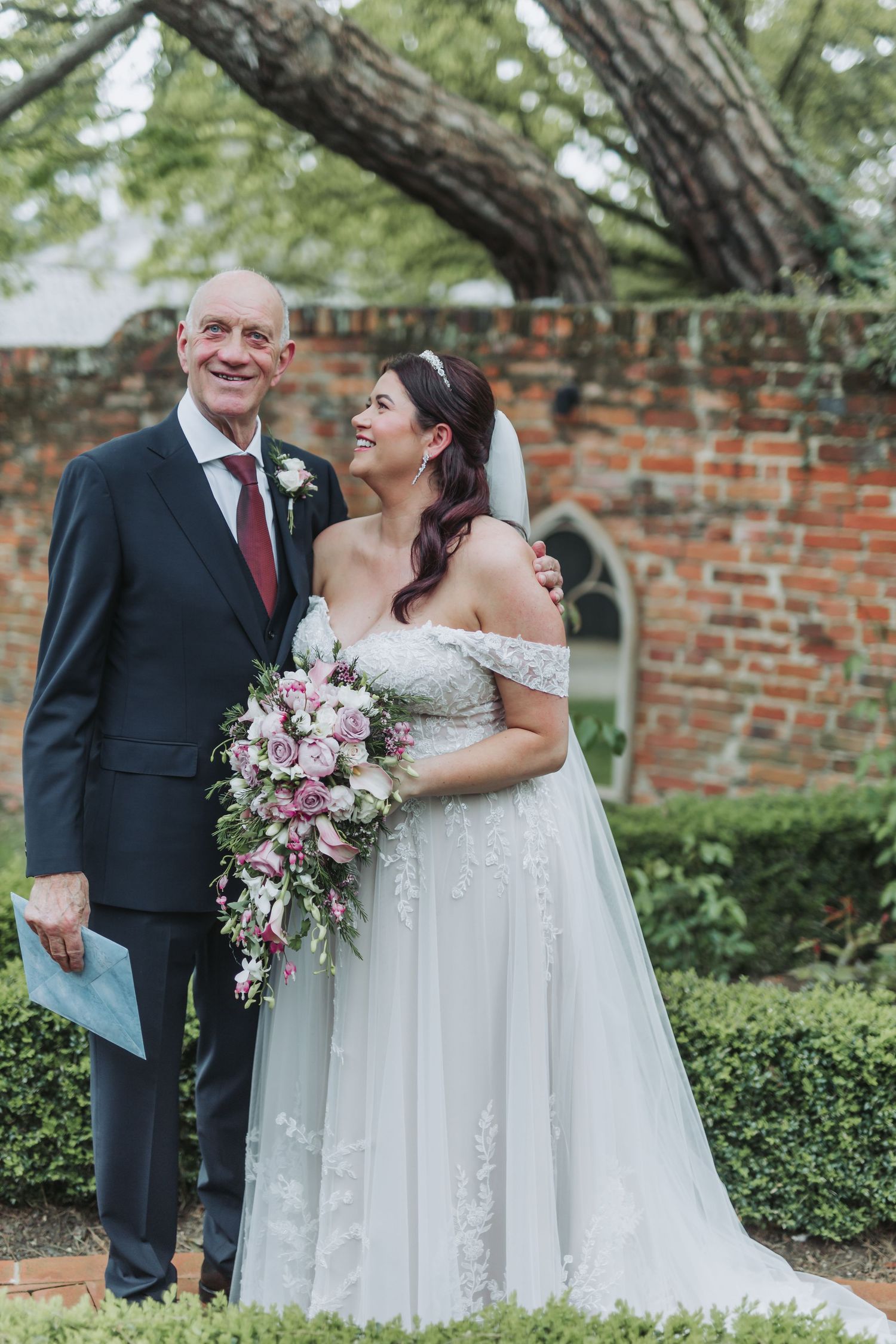 A couple shares a joyful moment in their wedding attire in front of a rustic brick wall and garden setting.
