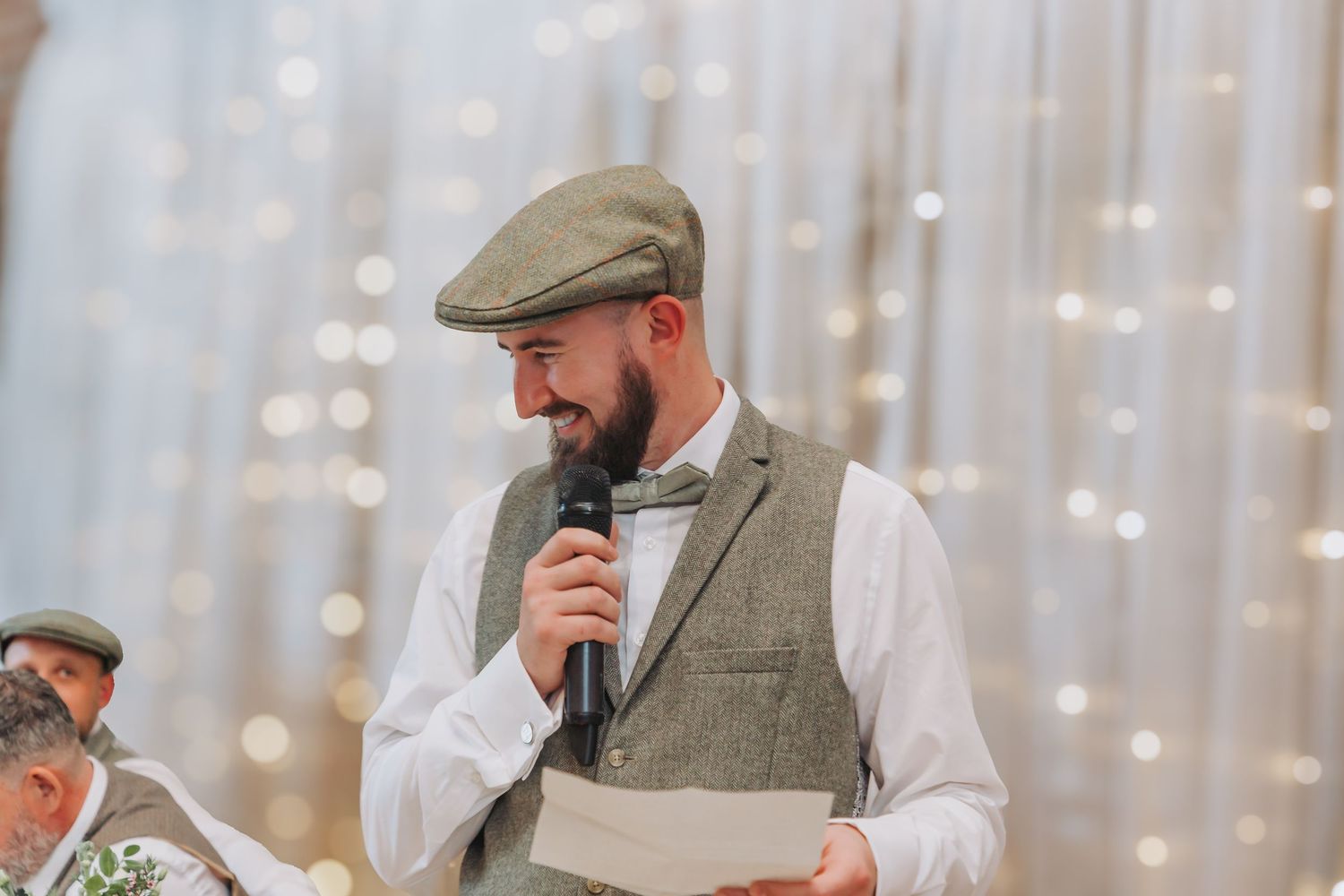 A groomsman in a tweed flat cap and gray suit gives a speech at a wedding reception with string lights in background.