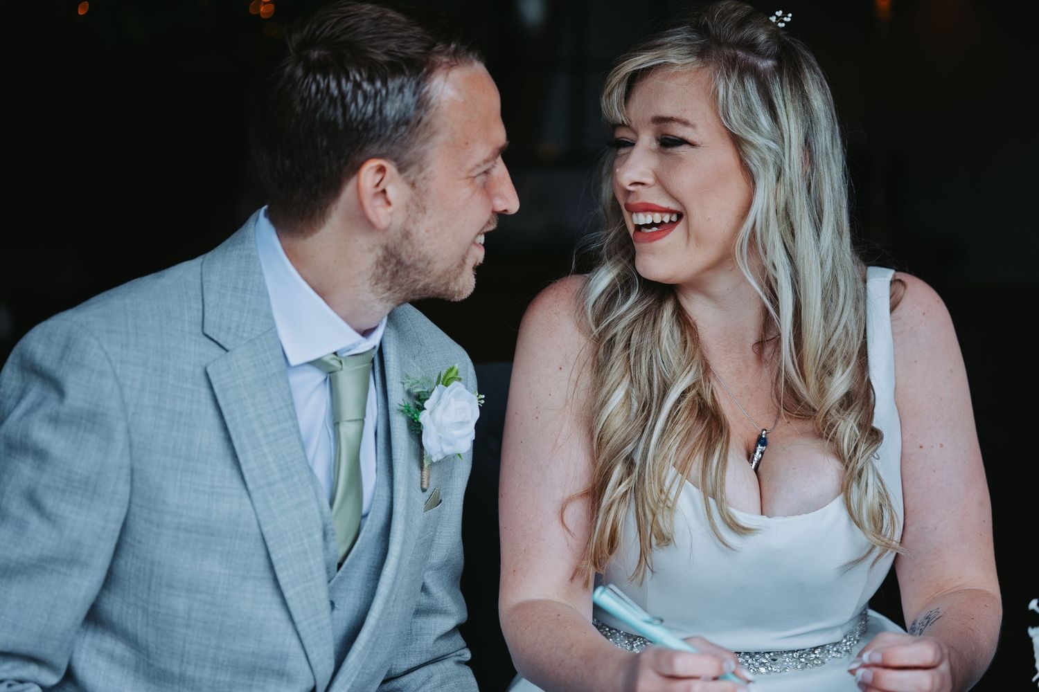 A laughing bride in a white dress and groom in a gray suit share a joyful moment together at their wedding reception.