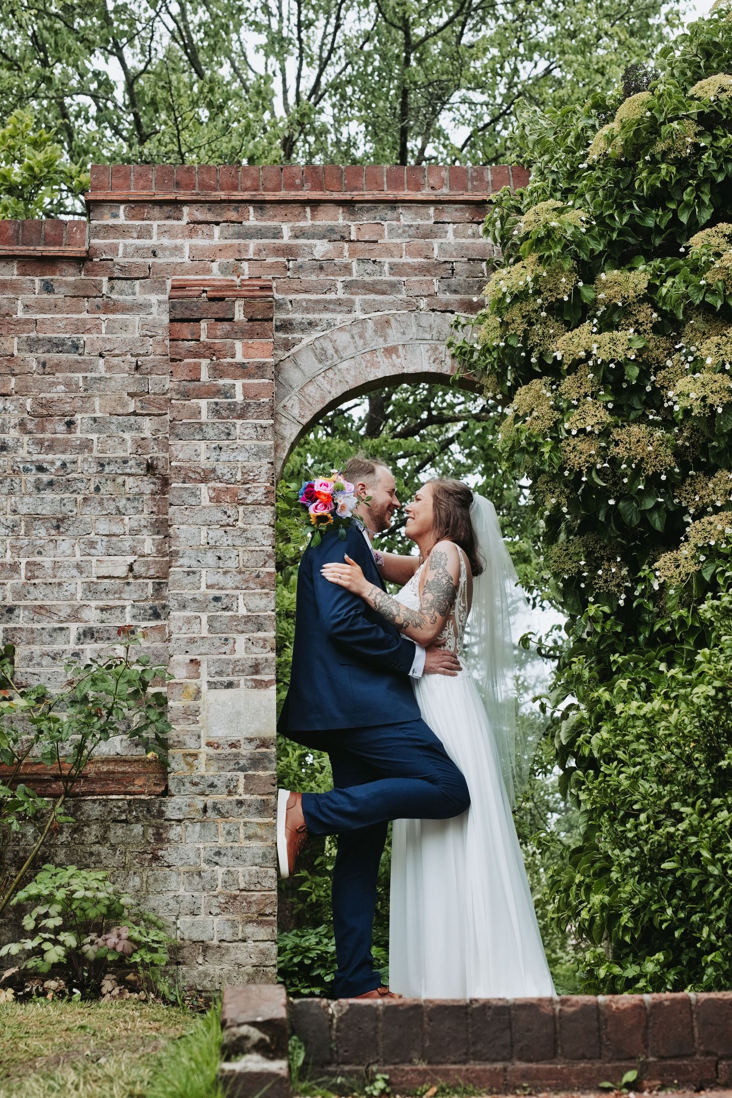 Bride and groom embrace under a rustic brick arch surrounded by lush greenery in a romantic garden wedding photo.