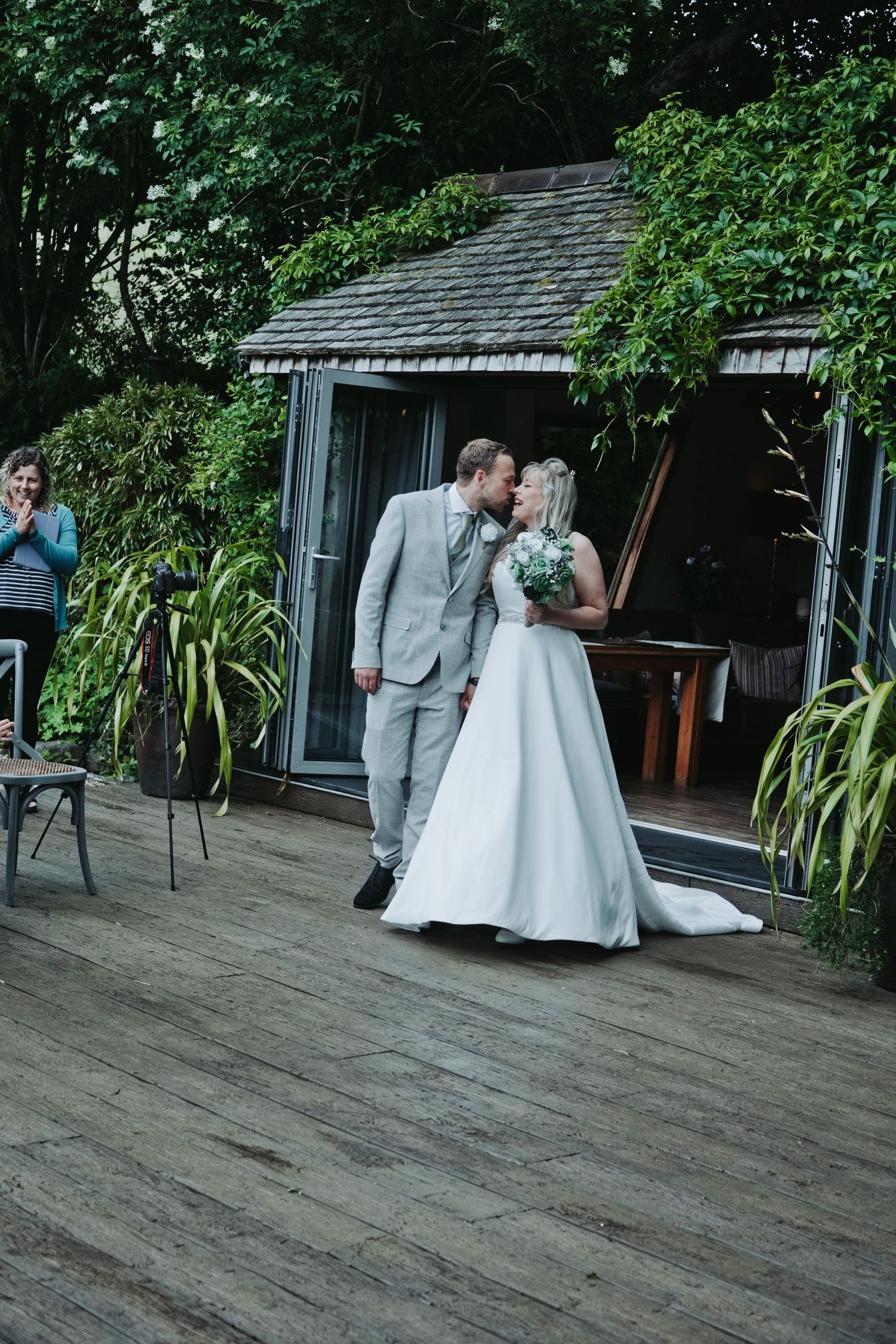 Bride and groom share first kiss on wooden deck surrounded by lush greenery at outdoor wedding ceremony.