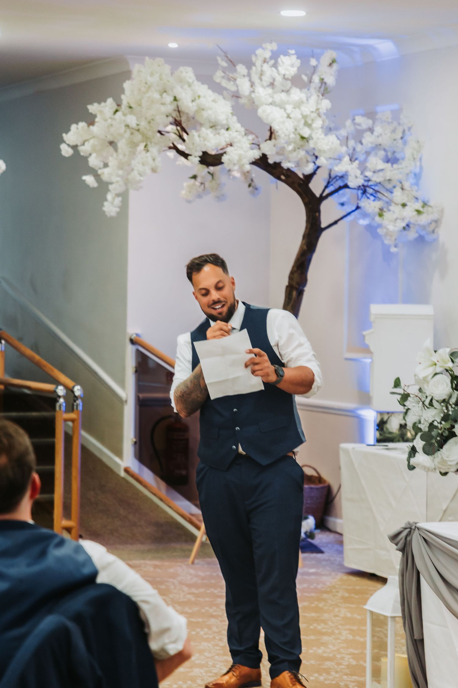A performer in a navy vest and white shirt stands in front of white cherry blossom trees at an indoor event venue.
