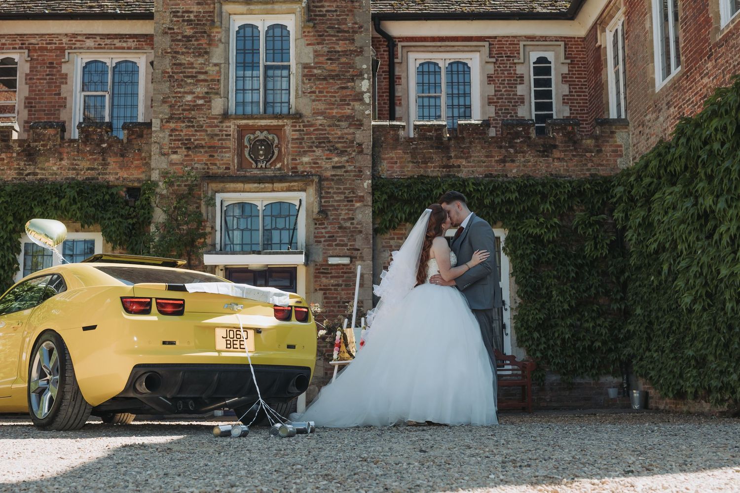 Elegant wedding couple poses with vintage yellow Chevrolet Camaro outside historic brick building.
