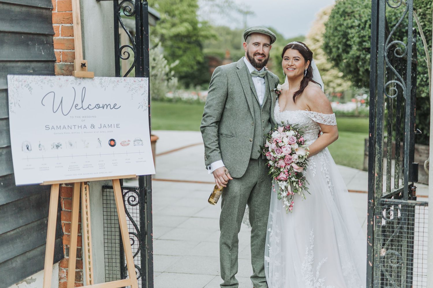 Wedding couple poses in front of rustic wooden doors with elegant floral decorations and welcome sign.