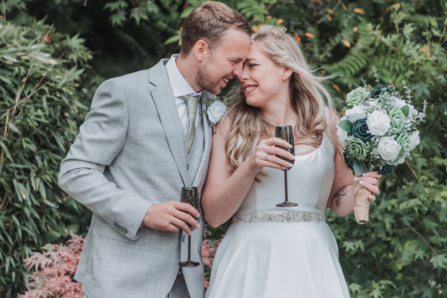 Happy newlyweds share a romantic moment, bride holding white bouquet and champagne flute, groom in grey suit outdoors.