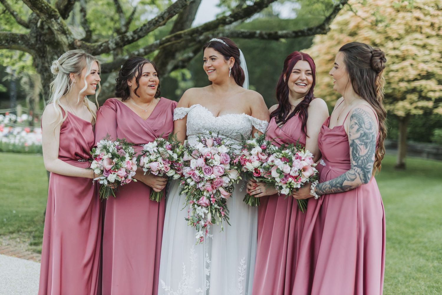 Bridesmaids in dusty rose dresses holding pink and white floral bouquets at an outdoor garden wedding ceremony.