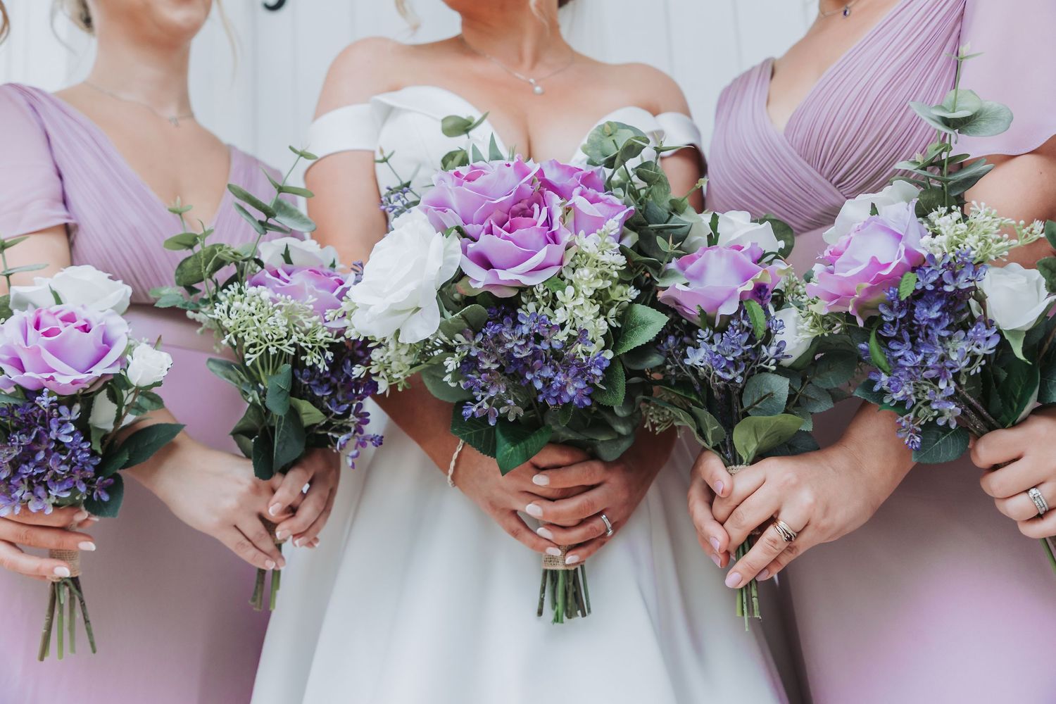Bridesmaids in blush pink dresses hold elegant bouquets of purple roses and white flowers at a wedding ceremony.