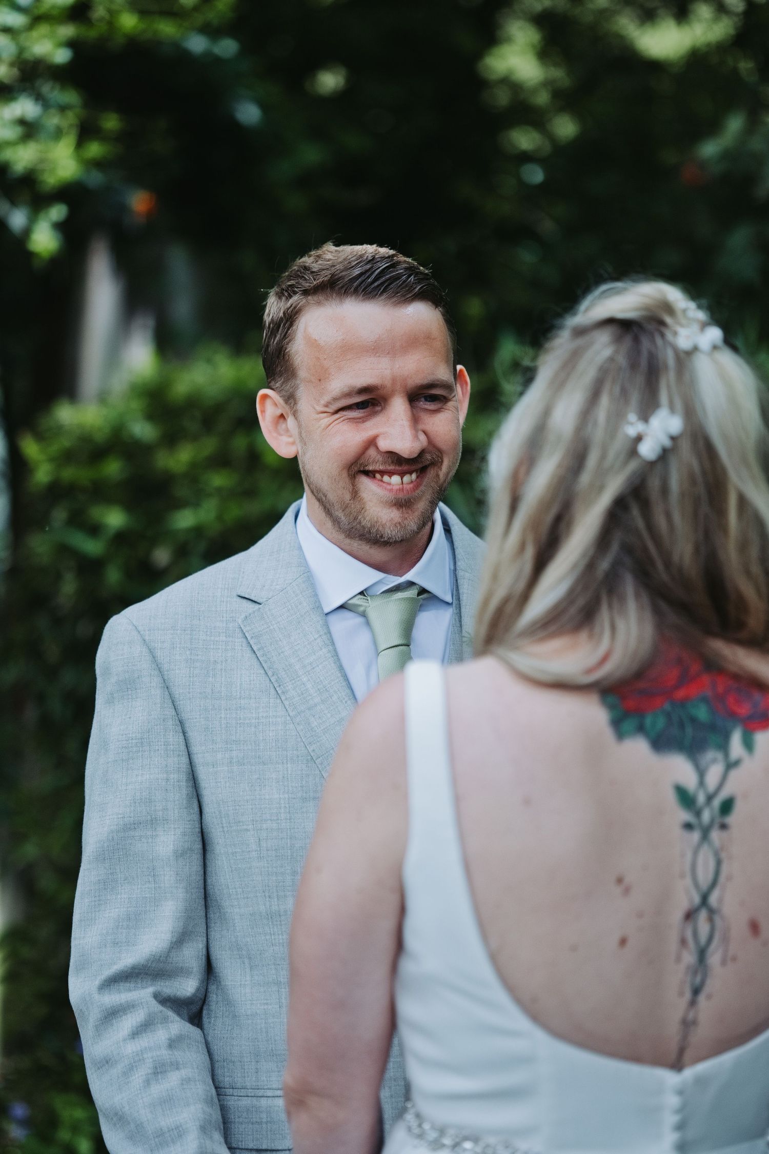 Smiling groom in gray suit faces tattooed bride during outdoor wedding ceremony surrounded by lush green trees.