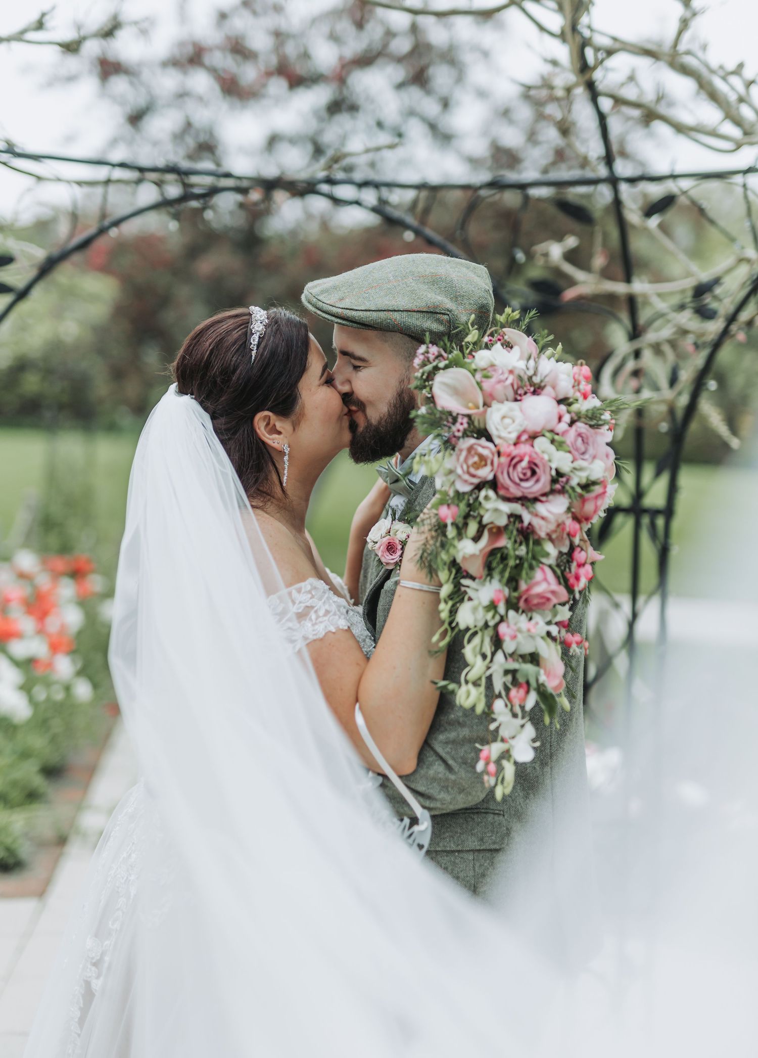A romantic wedding couple shares tender kisses behind pink orchid bouquets in a dreamy outdoor garden setting.