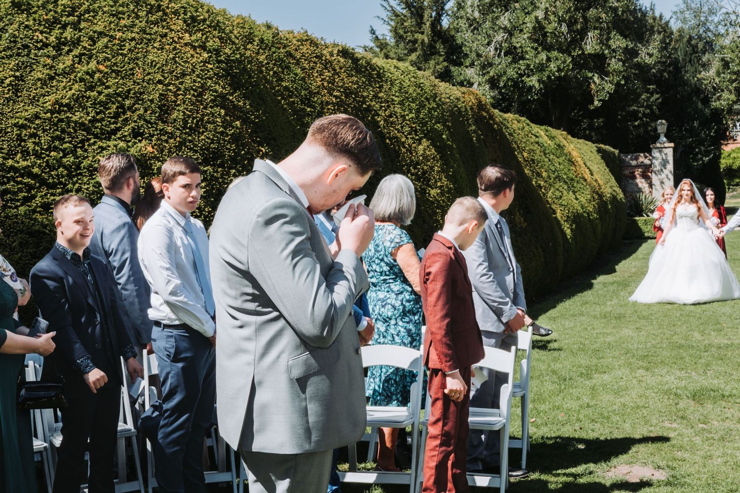 An emotional moment during an outdoor garden wedding ceremony with guests seated on white chairs and greenery in background.
