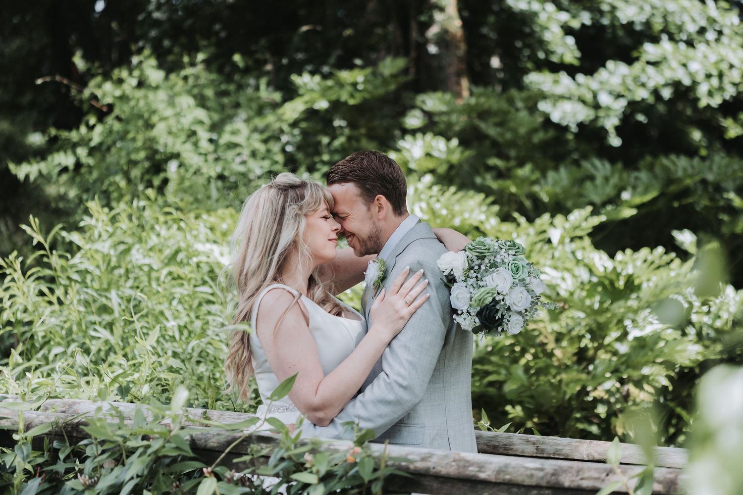 A couple shares a romantic moment surrounded by lush greenery, with the bride holding a white bouquet.