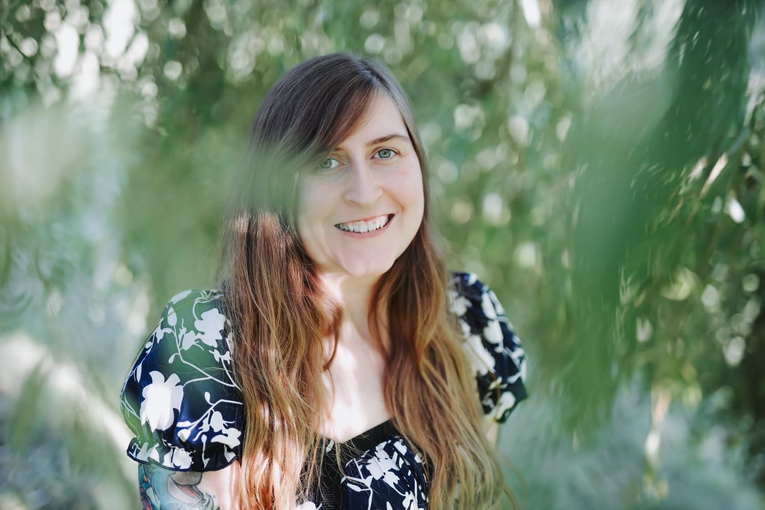 Smiling young woman with long brown hair wearing a floral dress, surrounded by lush green foliage outdoors.