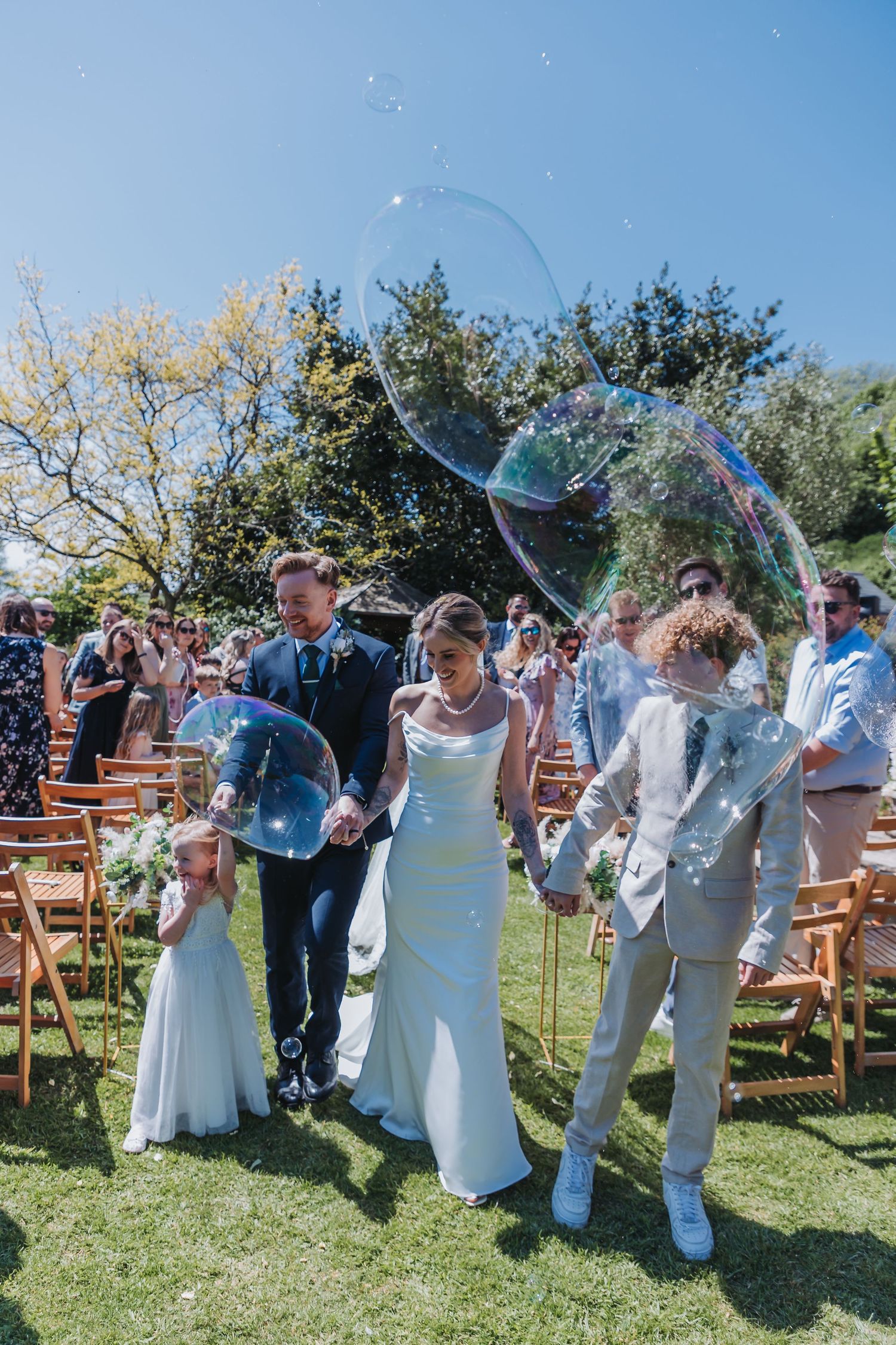 Wedding guests playing with giant bubbles in a sunny outdoor garden ceremony with wooden chairs.