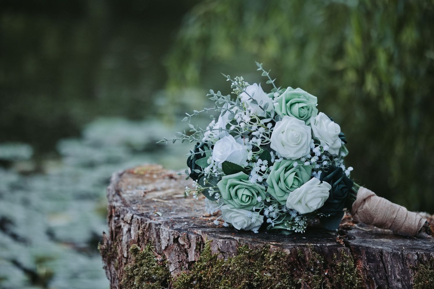 A bridal bouquet of mint green and white roses resting on a mossy tree stump near a serene pond.