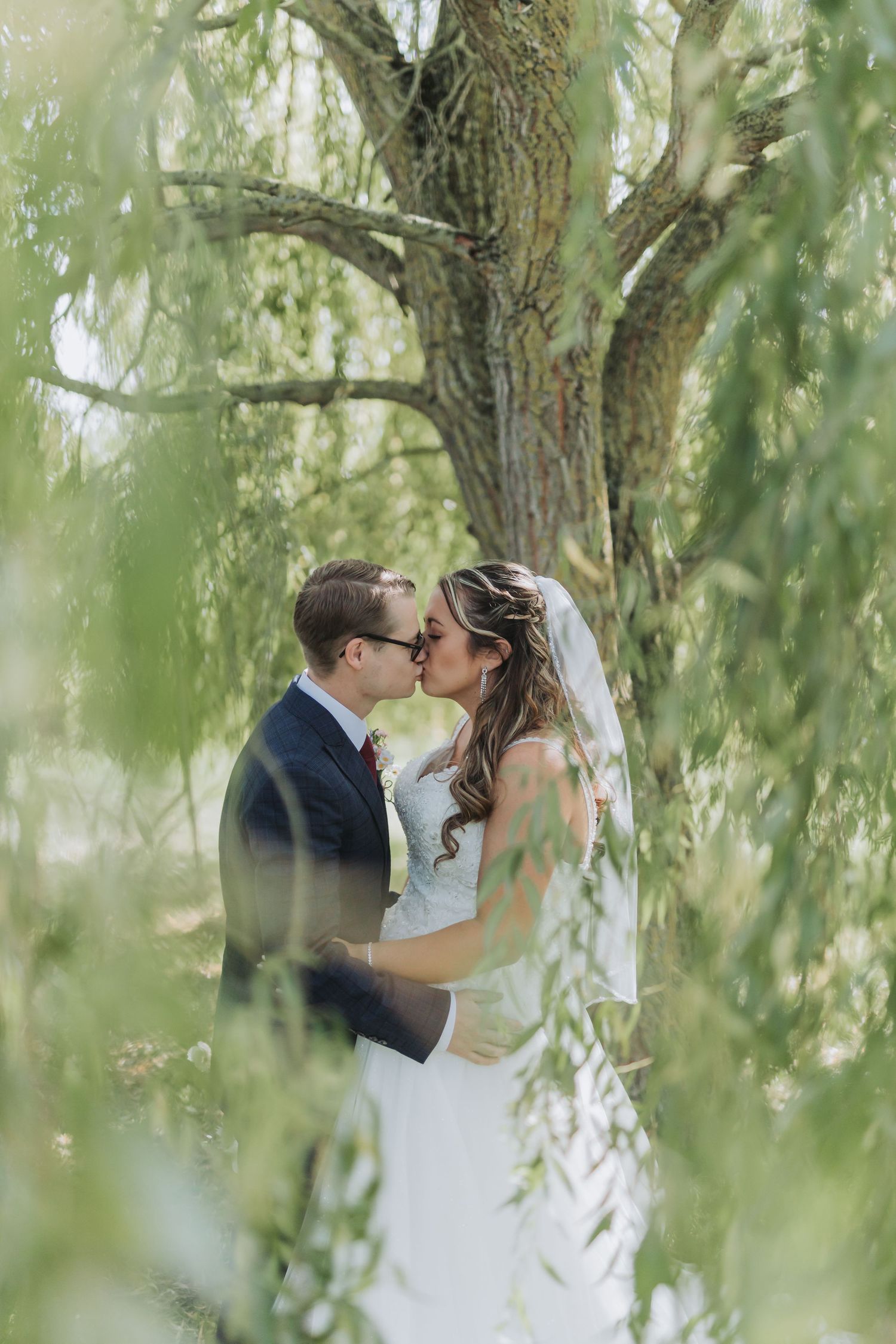 Romantic newlyweds share intimate moments beneath cascading willow tree branches in a dreamy outdoor setting.