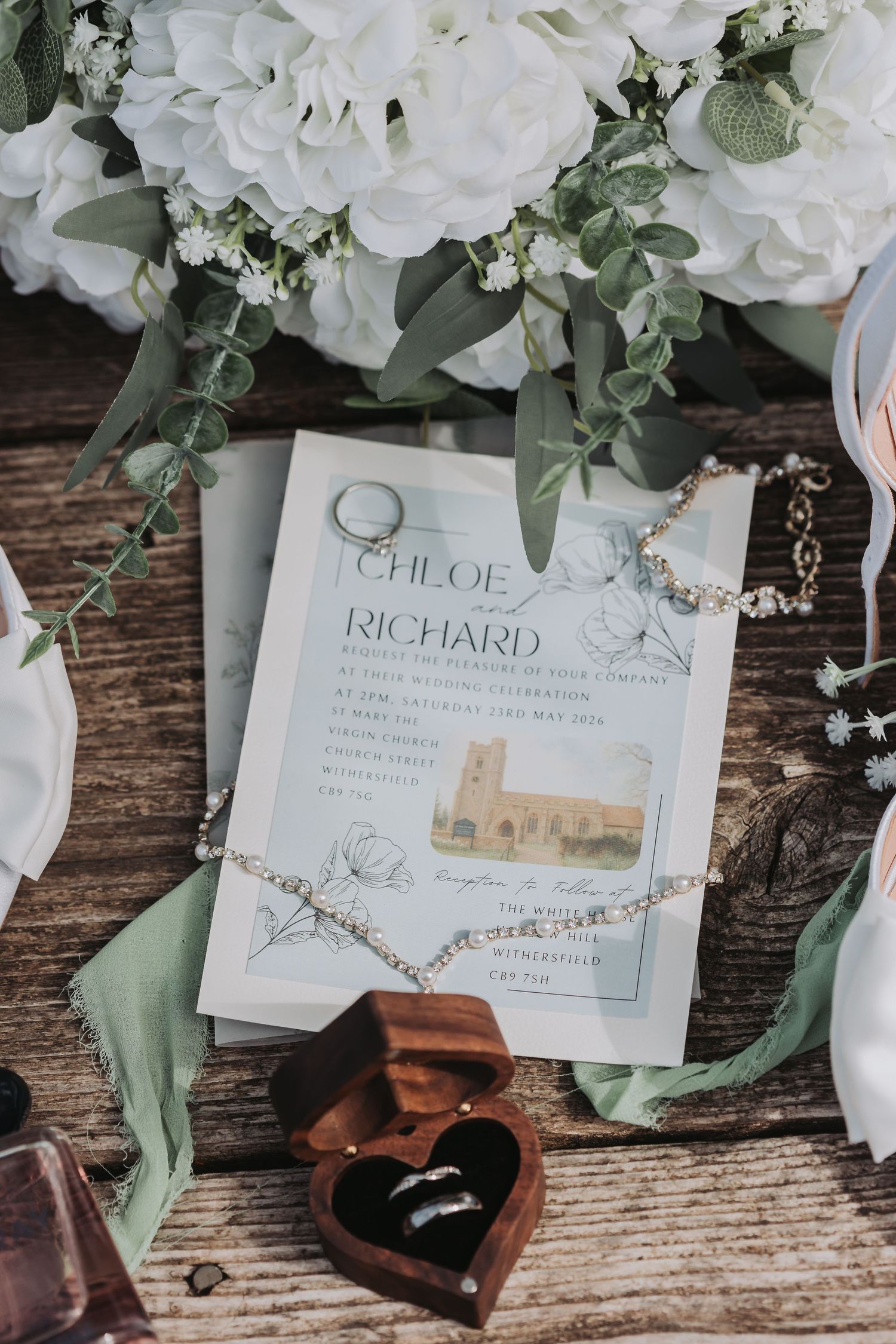Wedding flat lay with Chloe and Richard invitation, floral bouquet, rings in heart box, and sage green ribbon on rustic wood.