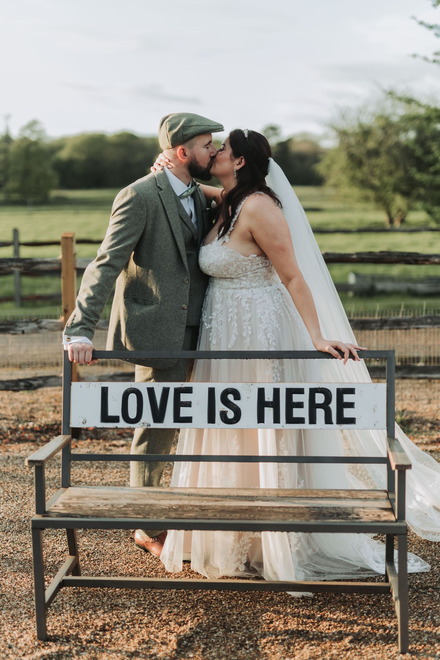 Newlyweds share intimate moments on rustic wooden bench with 'LOVE IS HERE' text in countryside setting.