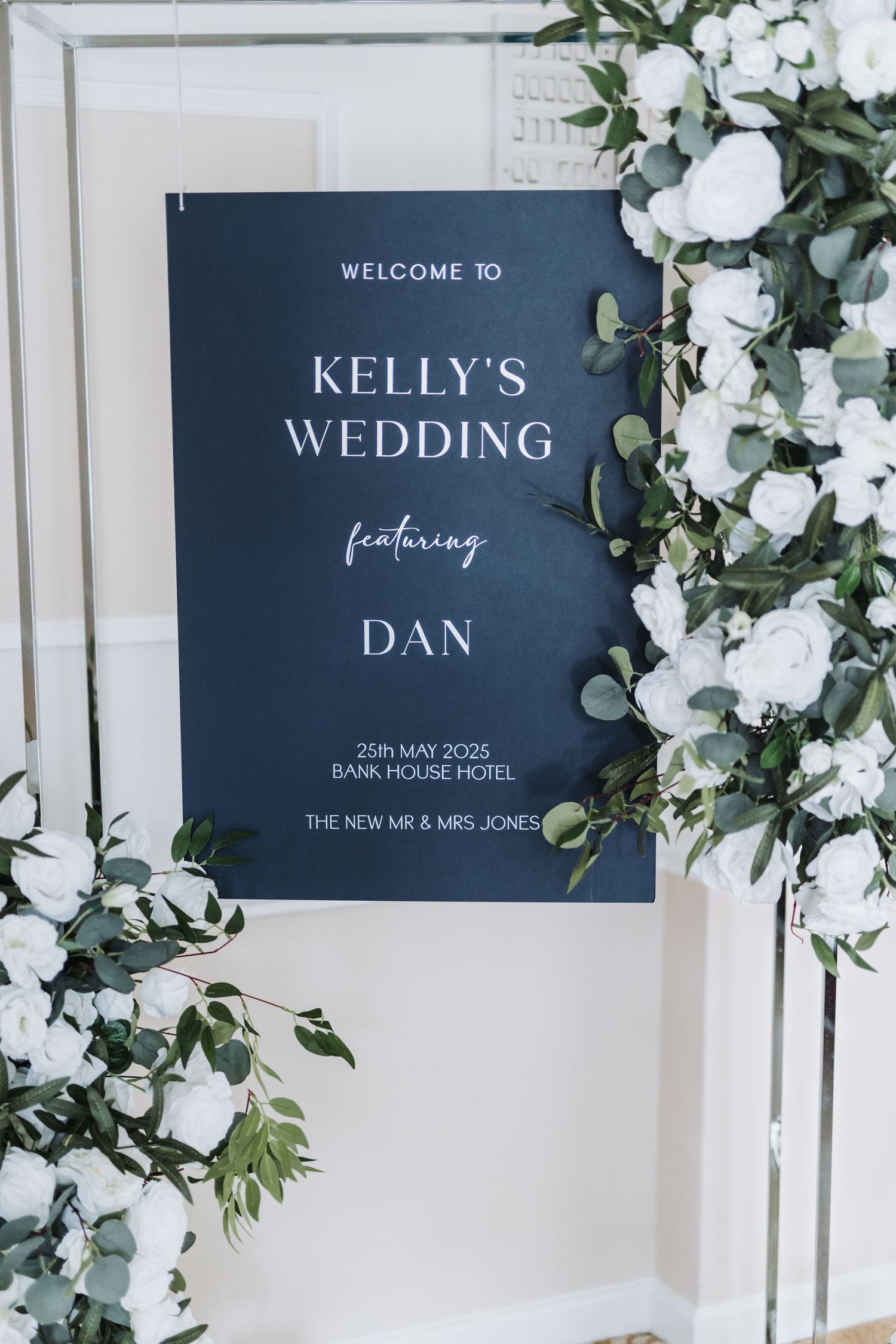 Modern navy blue wedding welcome signs with white text and greenery floral decorations against a light wall.