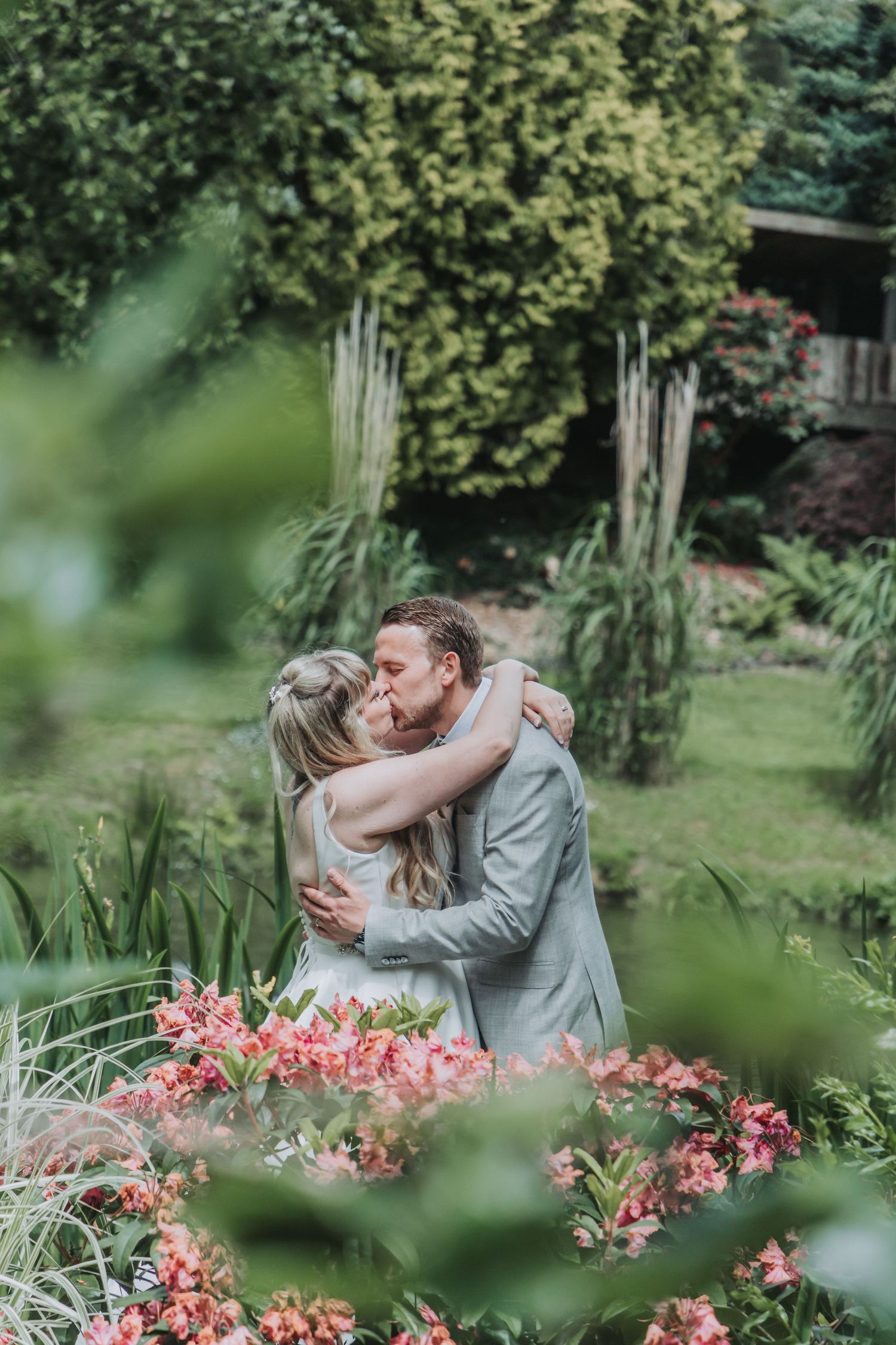 A couple shares a romantic kiss surrounded by lush greenery and pink flowers in a garden wedding setting.