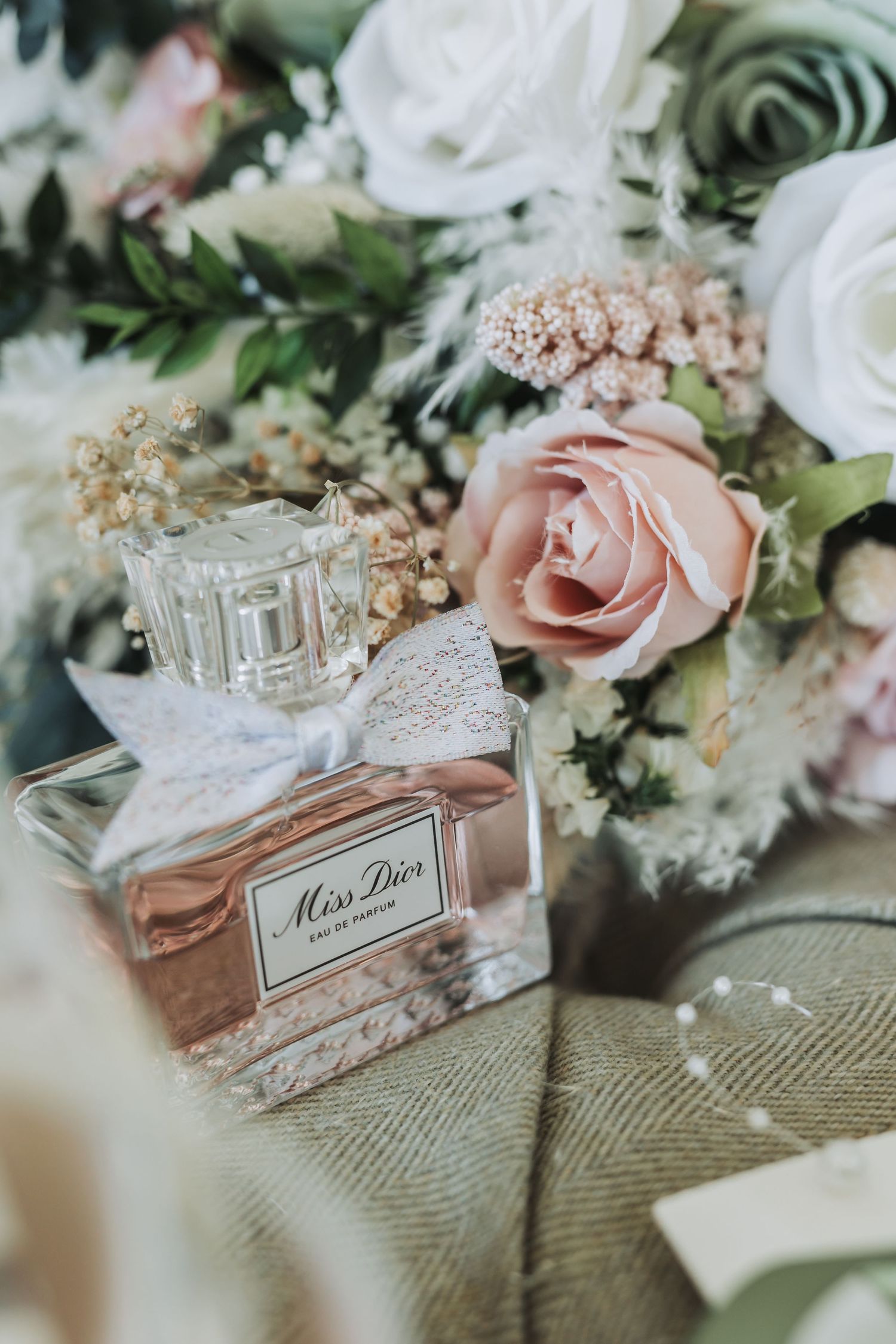 Miss Dior perfume bottle surrounded by delicate pink roses and baby's breath flowers on a textured fabric surface.