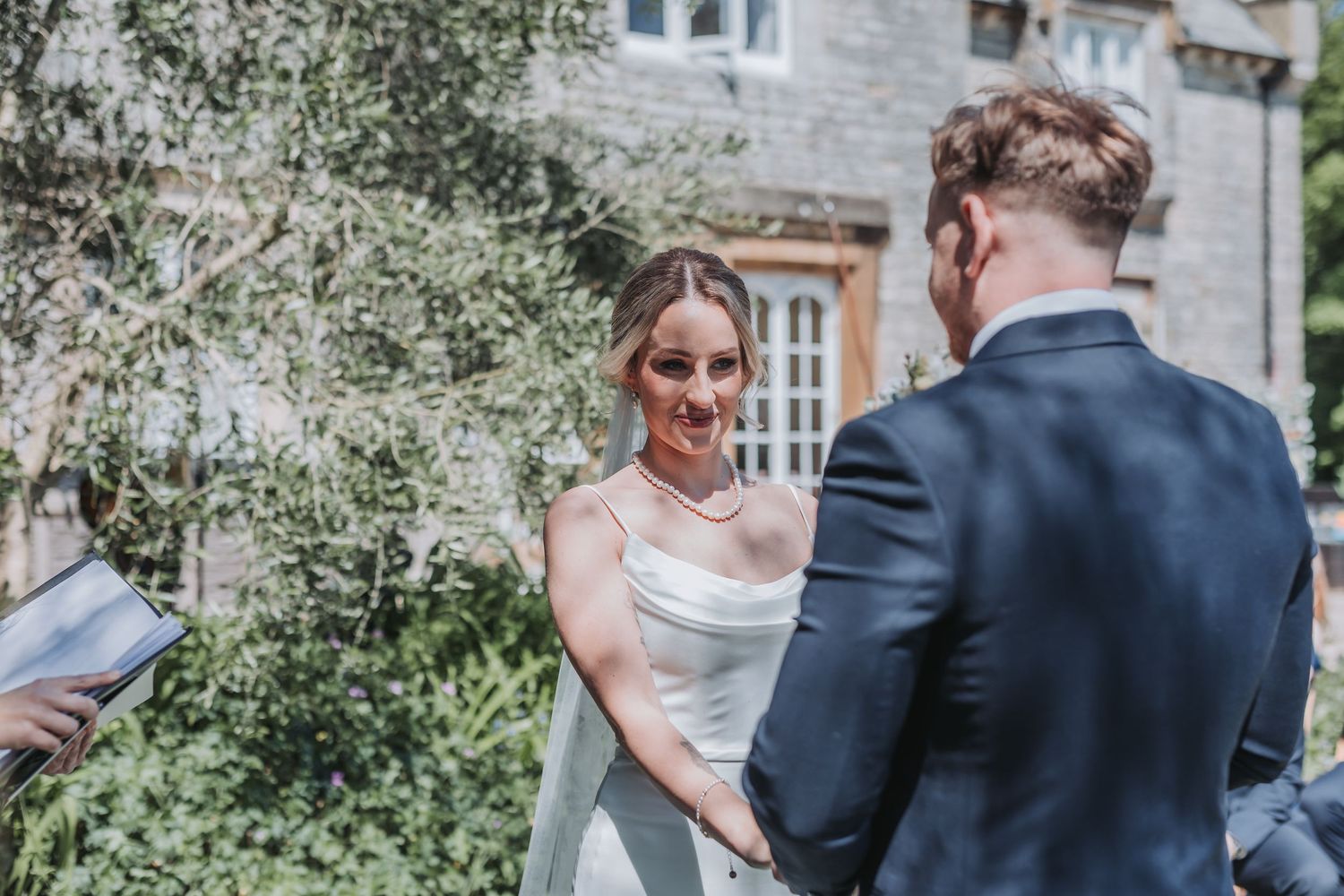 A wedding couple shares an intimate moment in a garden setting with stone architecture in the background.