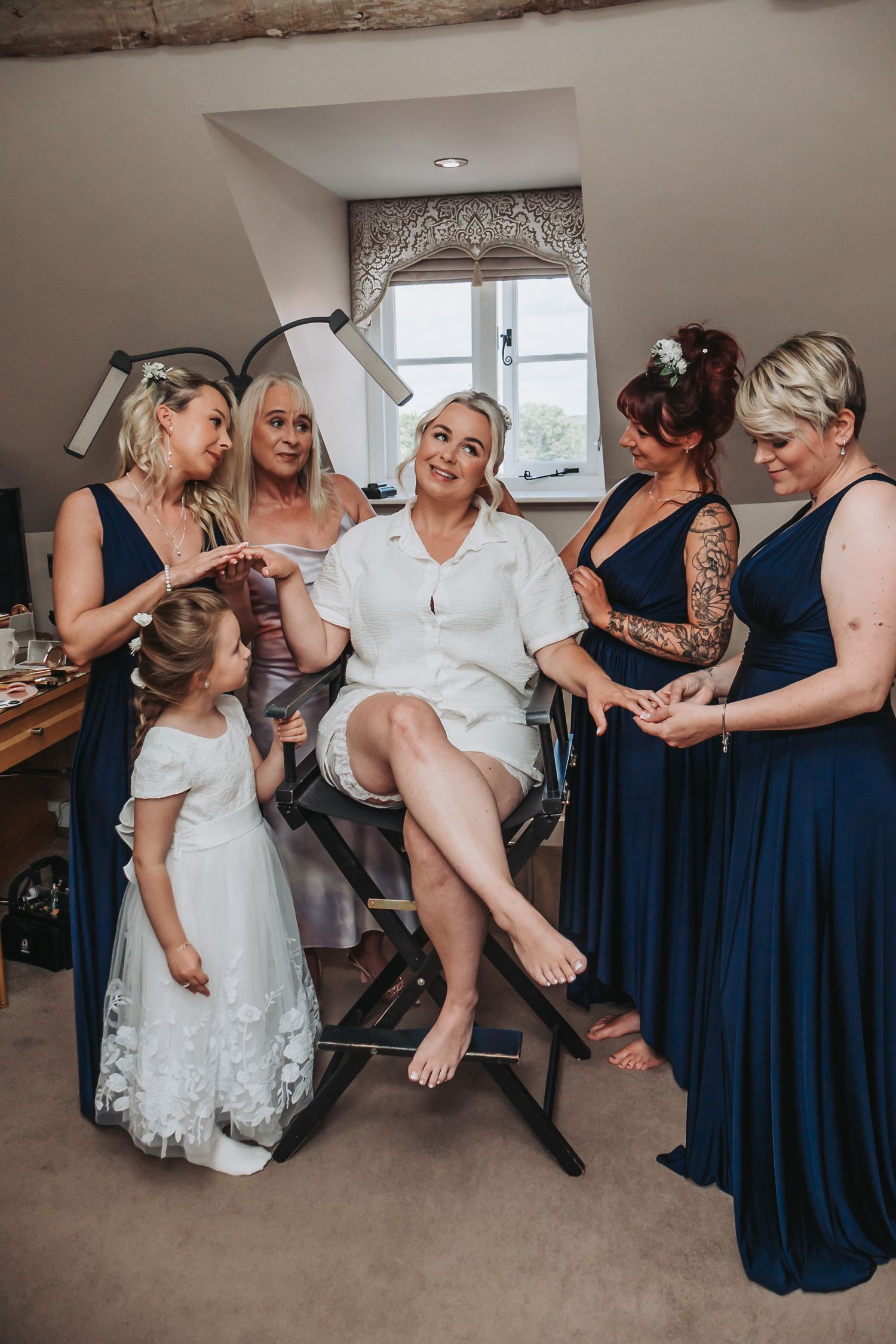 Wedding party helps bride put on a garter while wearing navy blue bridesmaid dresses and white bridal gown.