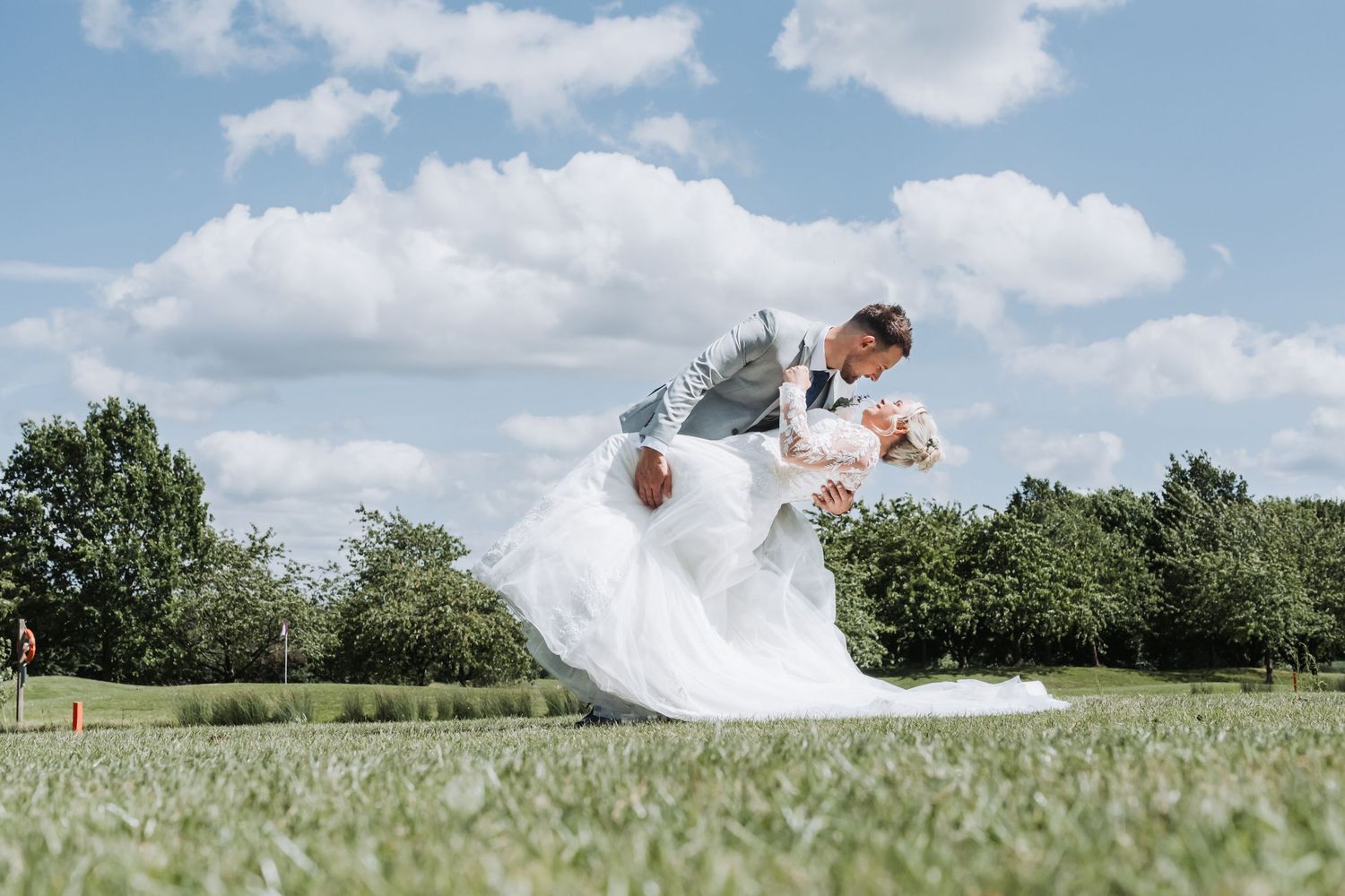 A bride and groom jump playfully in a lush green field under blue summer skies.
