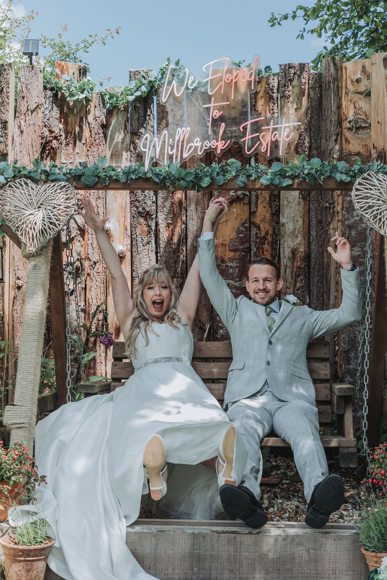 Joyful bride and groom celebrate on a swing at Millbrook Estate beneath a glowing neon wedding sign.