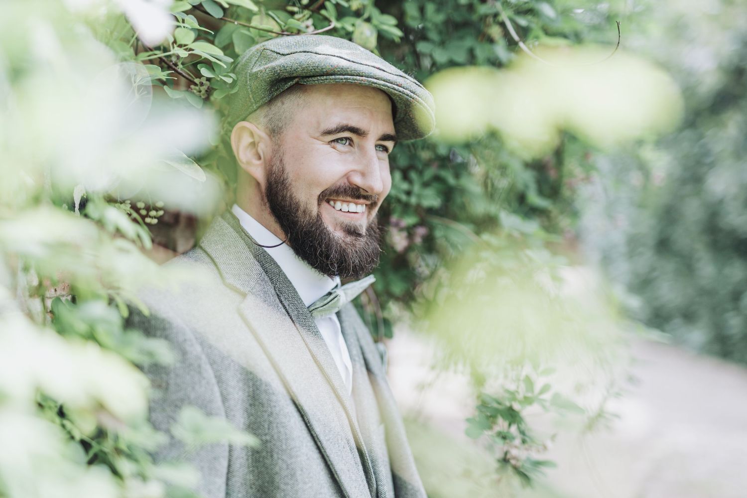 A person in a light gray suit and hat stands outdoors among green foliage in a natural setting.